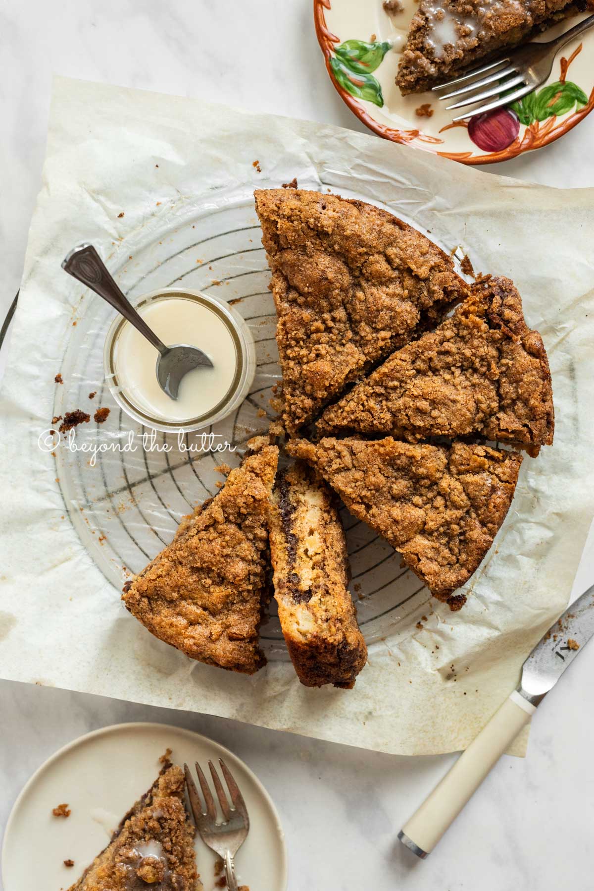 Slices of apple coffee cake on top of a parchment paper lined wire cooling rack with a small jar of simple vanilla glaze next to it | All Images © Luyiz™