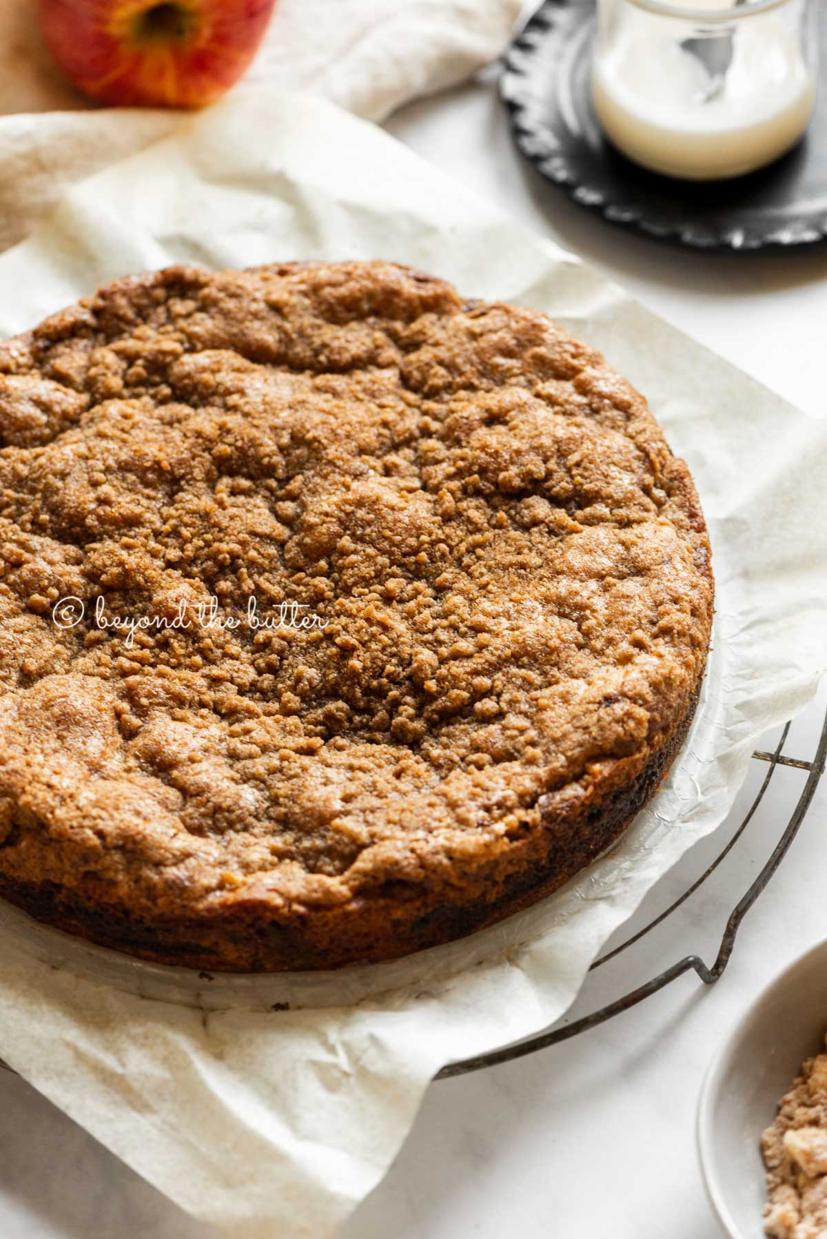 Cinnamon apple coffee cake colling on a parchment paper lined cooling rack with a glass of a simple vanilla glaze off to the side | All Images © Luyiz™