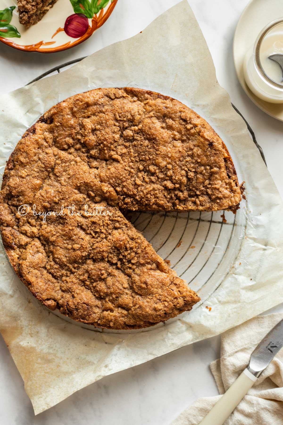 Apple coffee cake on a parchment paper lined wire cooling rack with a slice removed and a simple vanilla glaze, dessert plates, and a knife to the side | All Images © Luyiz™