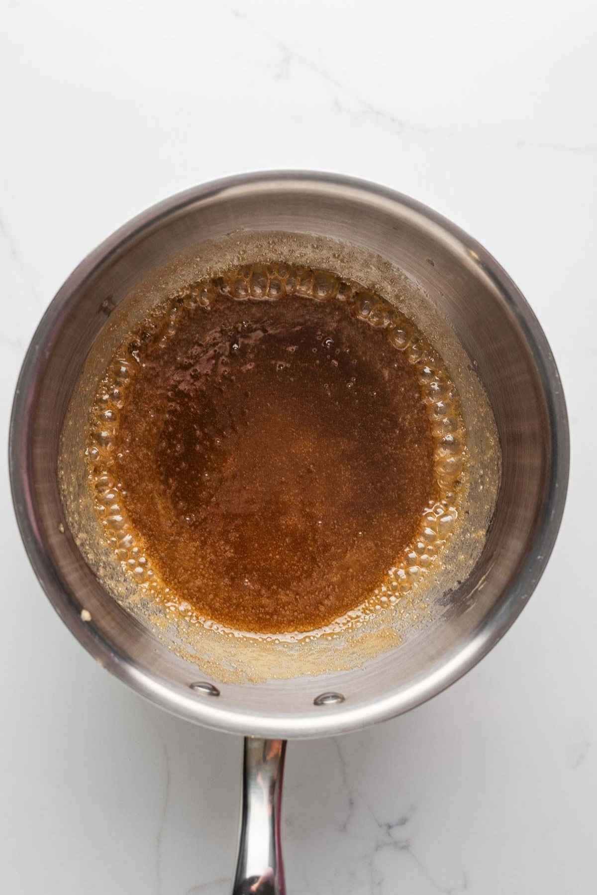 Brown sugar, butter, and heavy cream blended together and heated to a boil in a medium saucepan on a light grey marbled background.