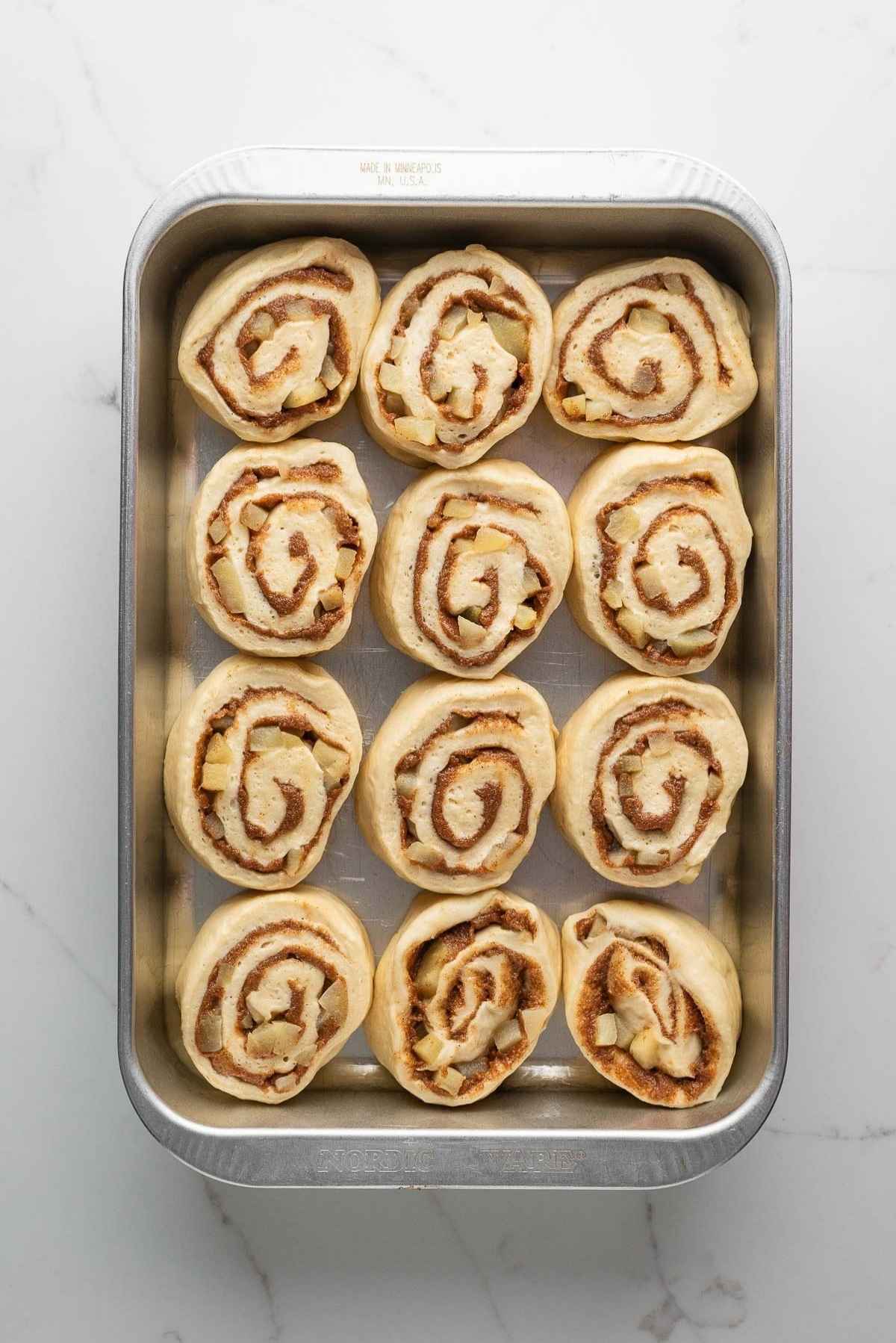 Apple cinnamon rolls sliced into 12 equally sized rolls and placed in a greased nine inch by thirteen inch metal pan on a light grey marbled background.