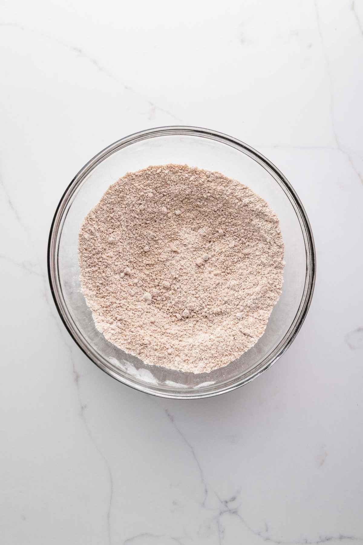 Whisked flour, brown sugar, and cinnamon in a medium glass mixing bowl on a light grey marbled background.