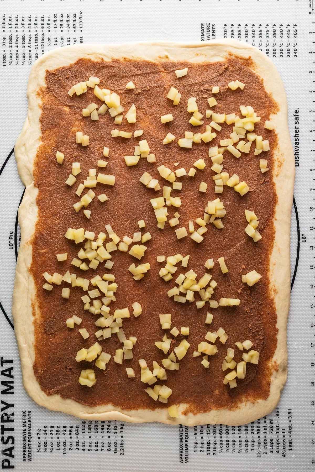 Brown sugar cinnamon filling spread across rolled out cinnamon roll dough and topped with chopped and softened apples on a silicone mat.