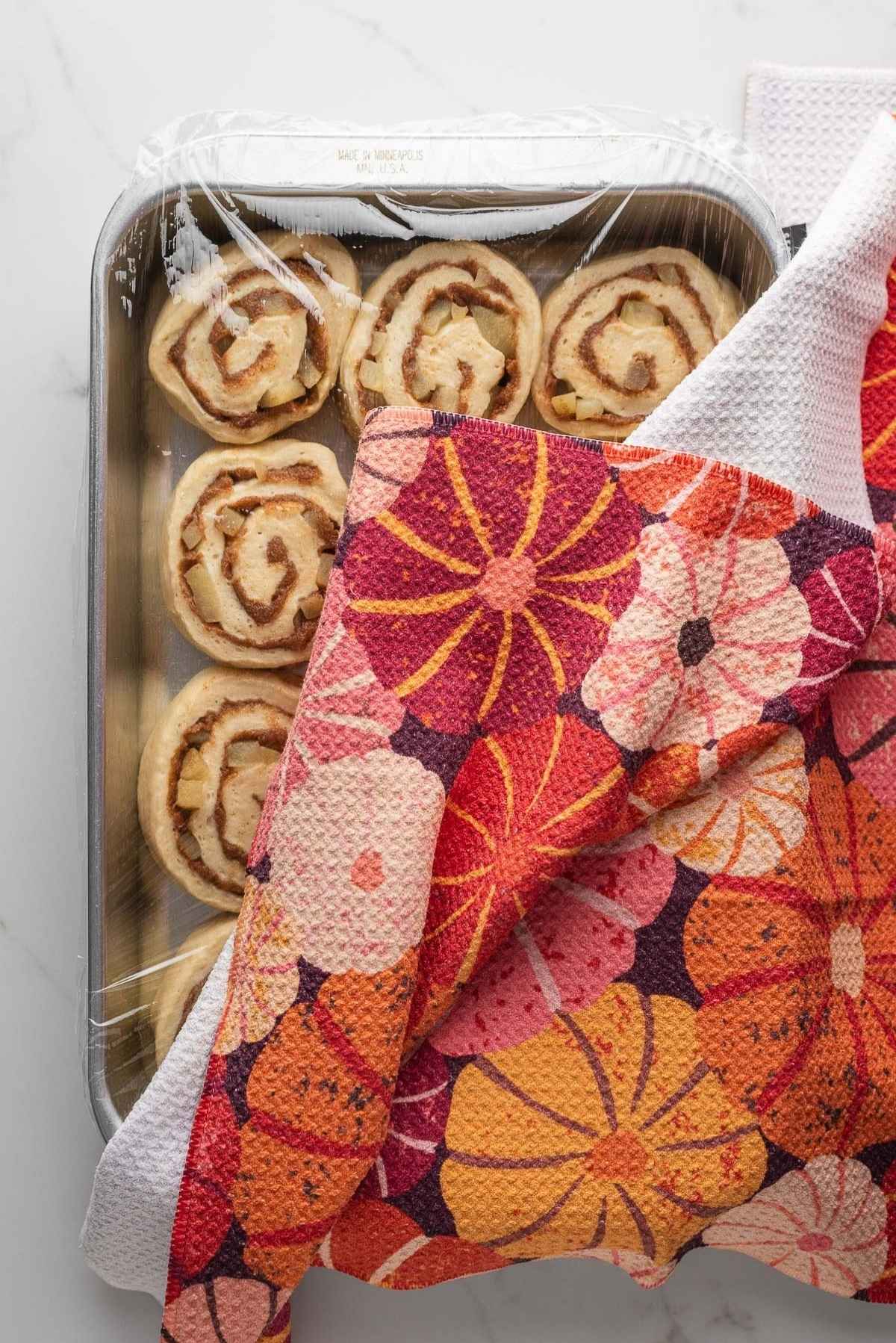 Apple cinnamon rolls loosely covered with plastic wrap and a clean kitchen towel on a light grey marbled background.