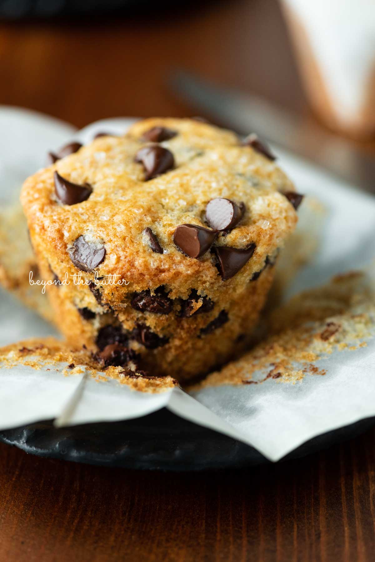 Unwrapped bakery style chocolate chip muffin on flattened muffin linerand black dessert plate | © Luyiz®