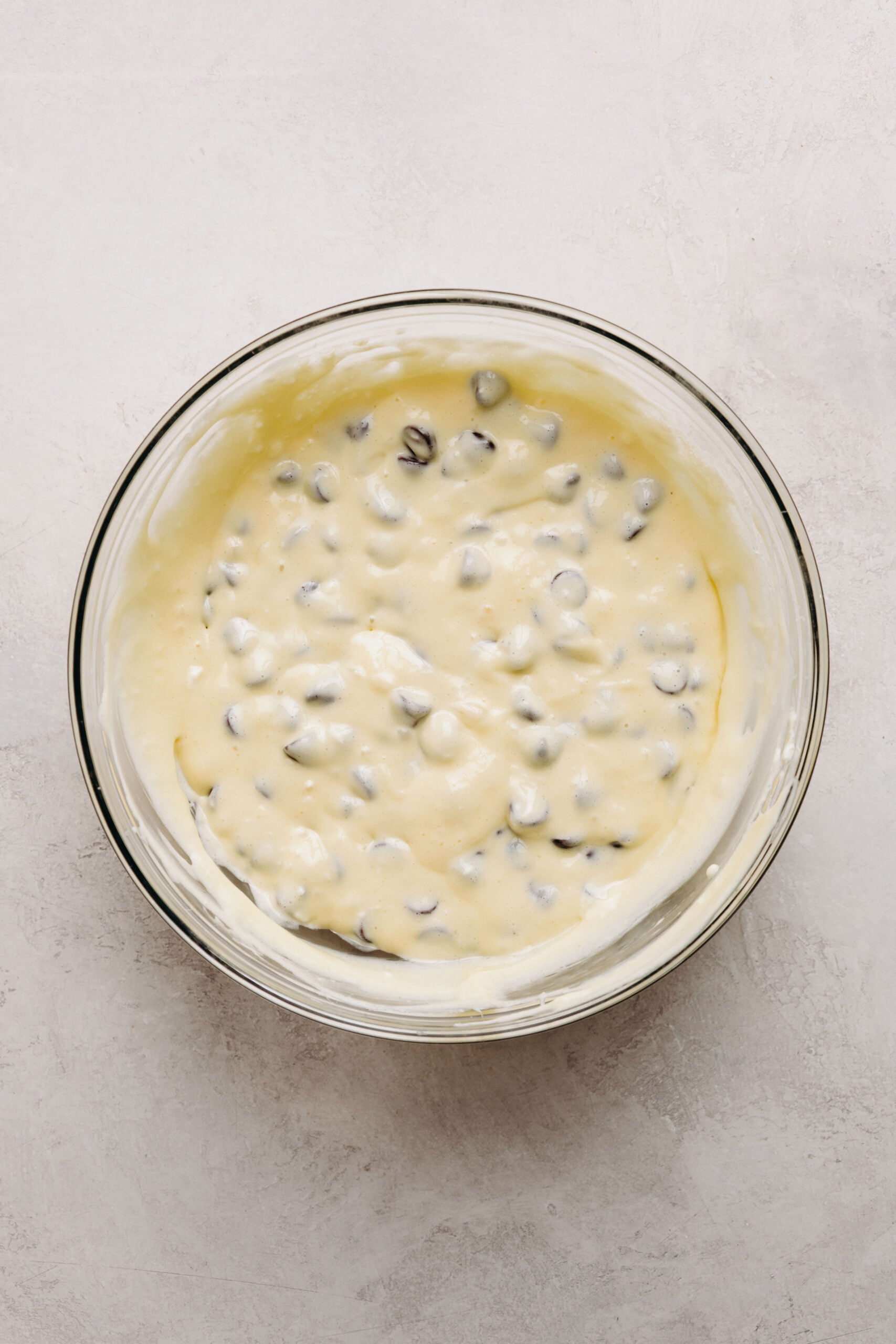 Chocolate chips folded into cream cheese filling in a glass mixing bowl on a white marbled background.
