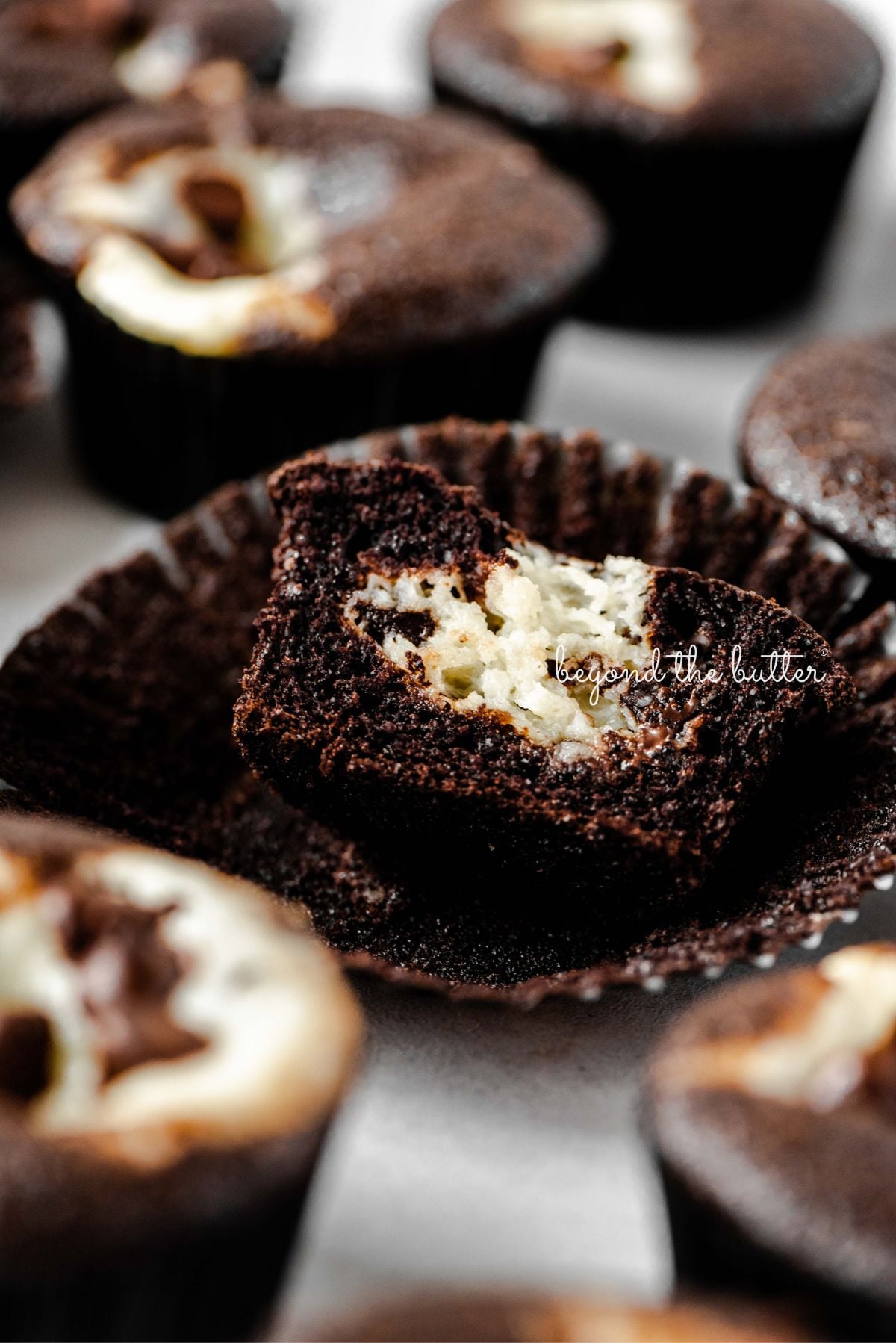 Cream cheese filled chocolate cupcakes topped with semi-sweet chocolate chips randomly placed on a light gray background with center cupcake unwrapped and sliced in half to see the cream cheese center.