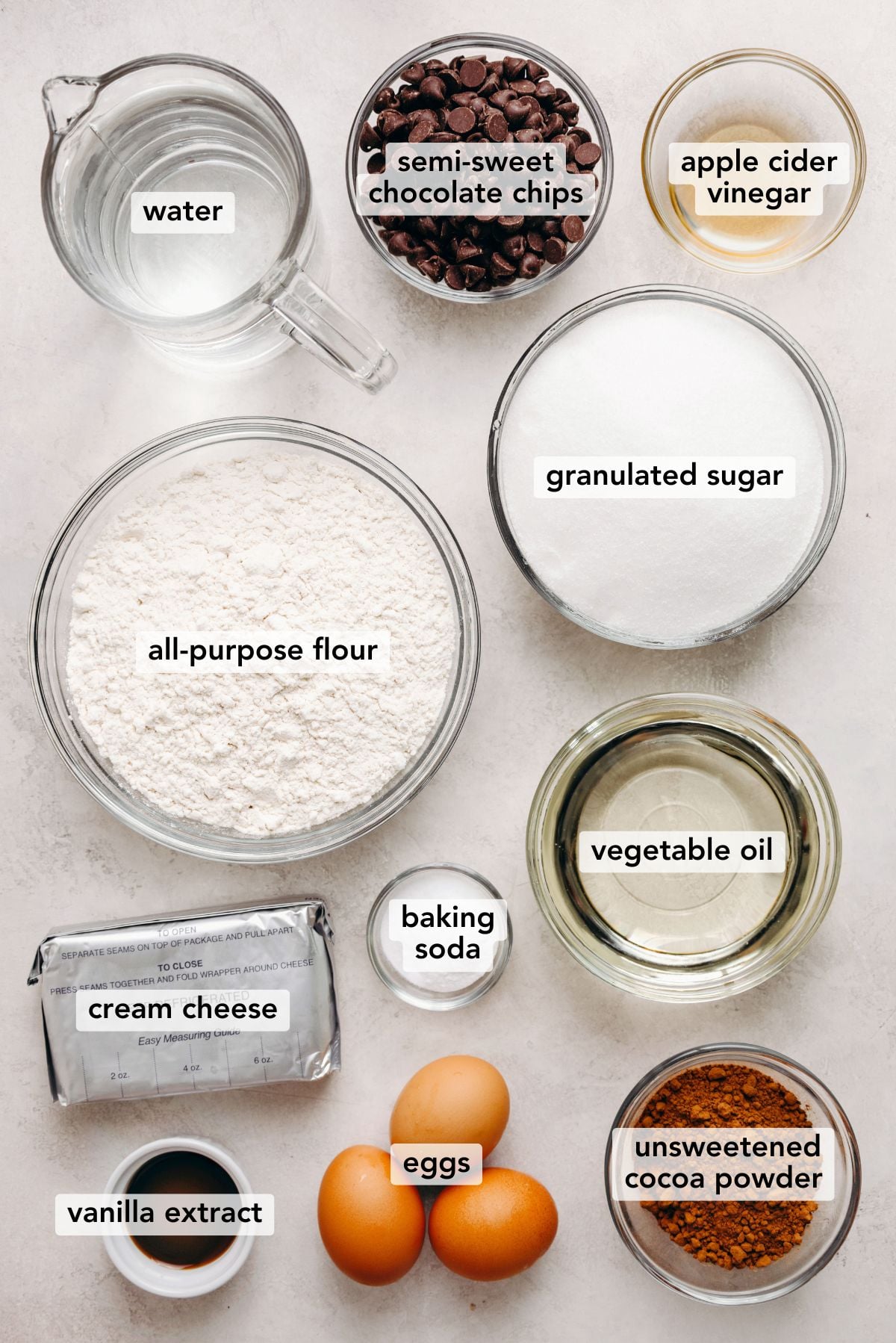 Ingredients in various size bowls labeled as water, semisweet chocolate chips, apple cider vinegar, all purpose flour, granulated sugar, cream cheese, baking soda, vegetable oil, vanilla extract, eggs, and unsweetened cocoa powder on a white marbled background.