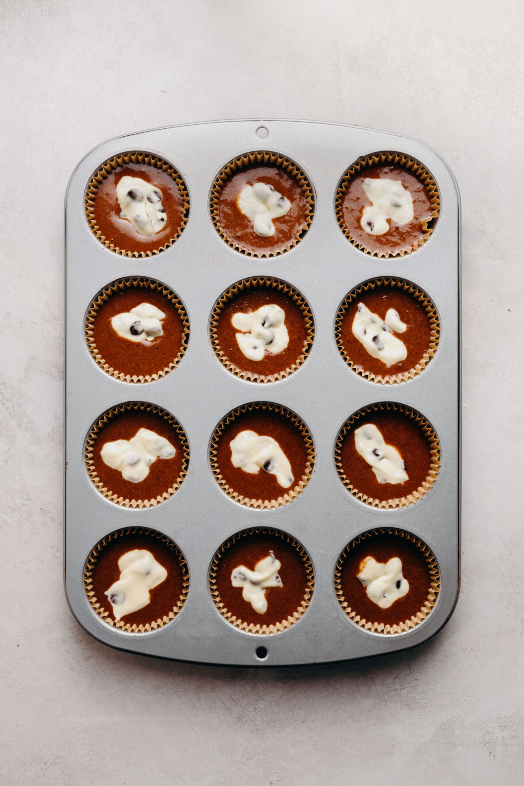Metal cupcake tin filled with chocolate cupcake batter topped with chocolate chip cream cheese filling on a white marbled background.