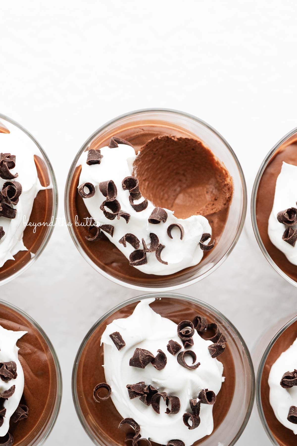 Six parfait goblets filled with chocolate mousse, whipped cream, and chocolate curls with a spoonful removed from the top center goblet on a white textured background.