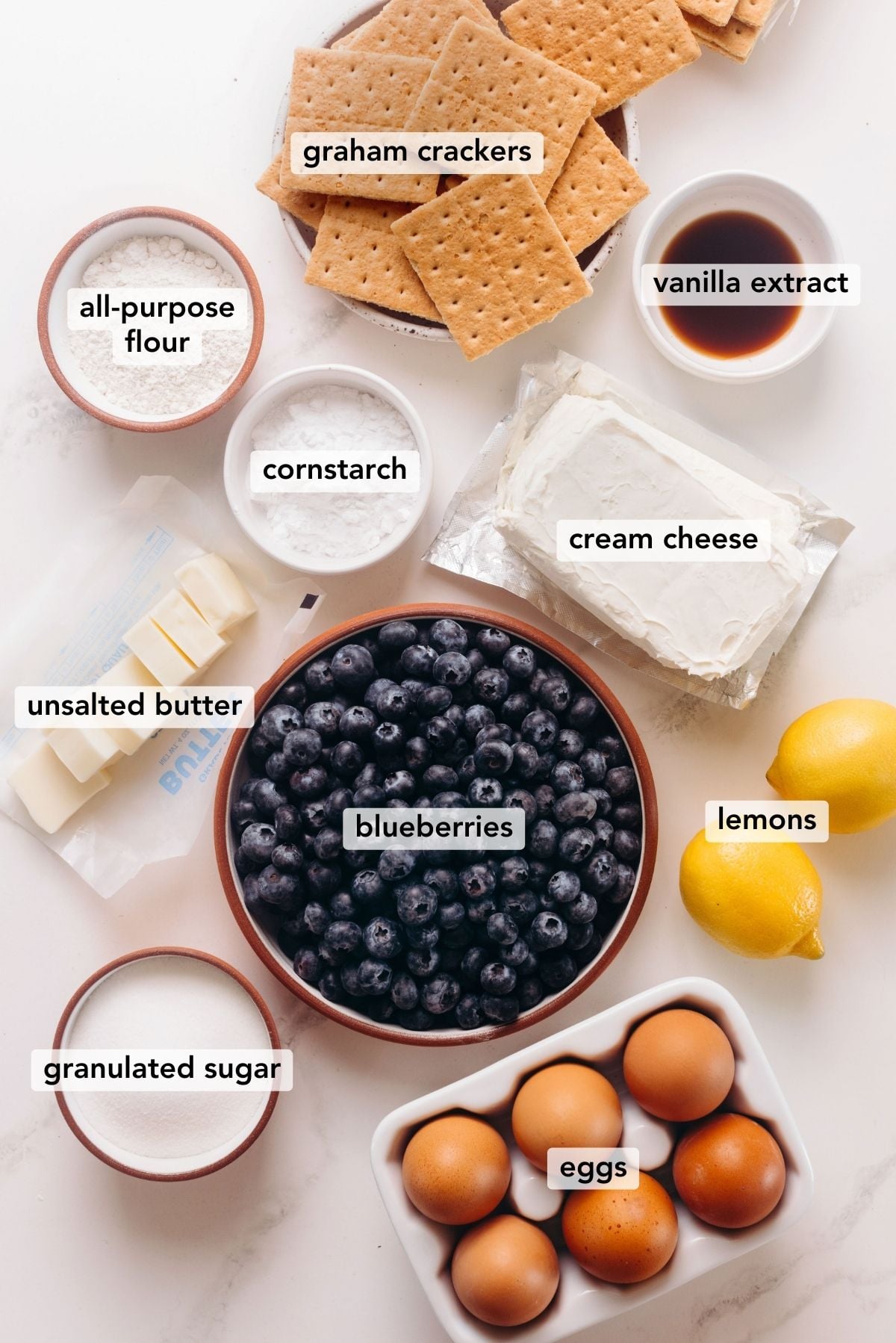 Ingredients in various size bowls labeled as blueberries, cornstarch, lemon juice, vanilla extract, granulated sugar, graham crackers, unsalted butter, cream cheese, eggs, and all-purpose flour on a white marbled background.