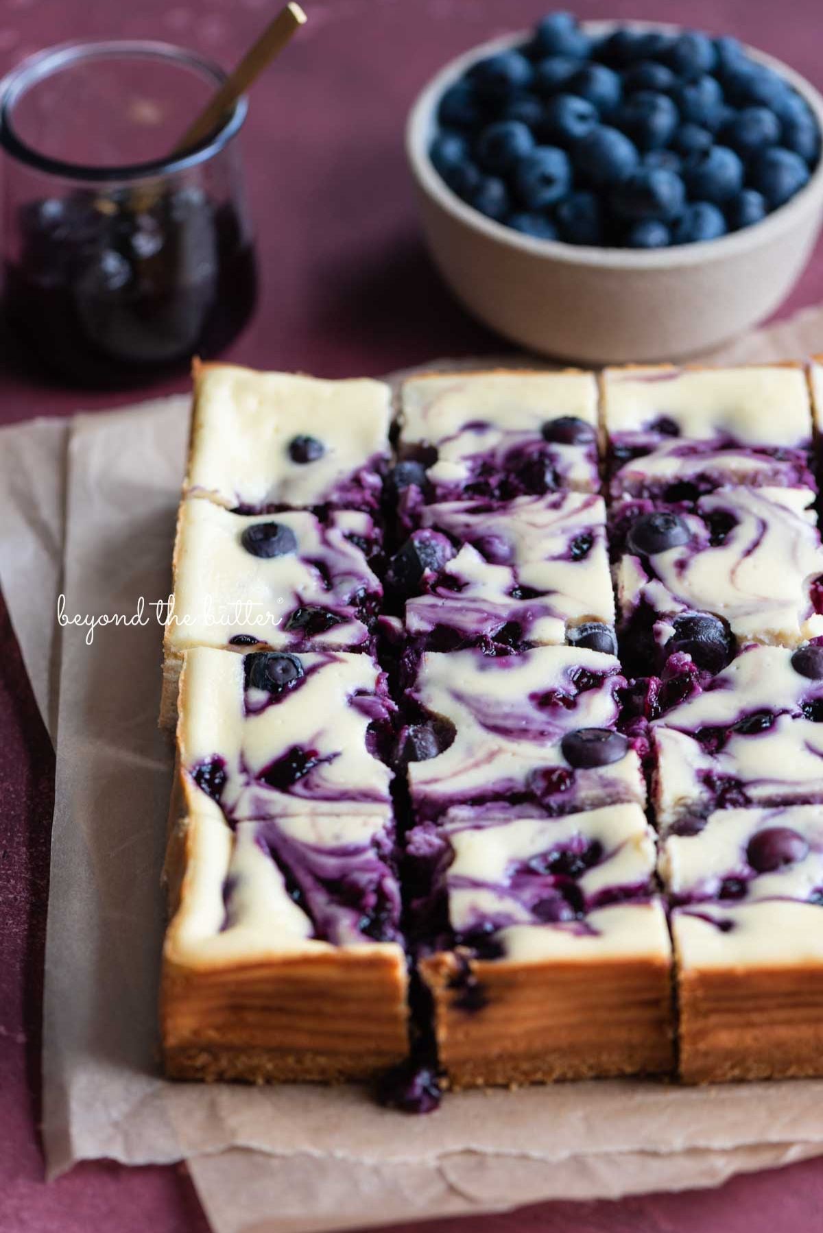 Slices of blueberry swirl cheesecake bars placed on two sheets of parchment paper with bowl of blueberry sauce and fresh blueberries to the side on pink background.