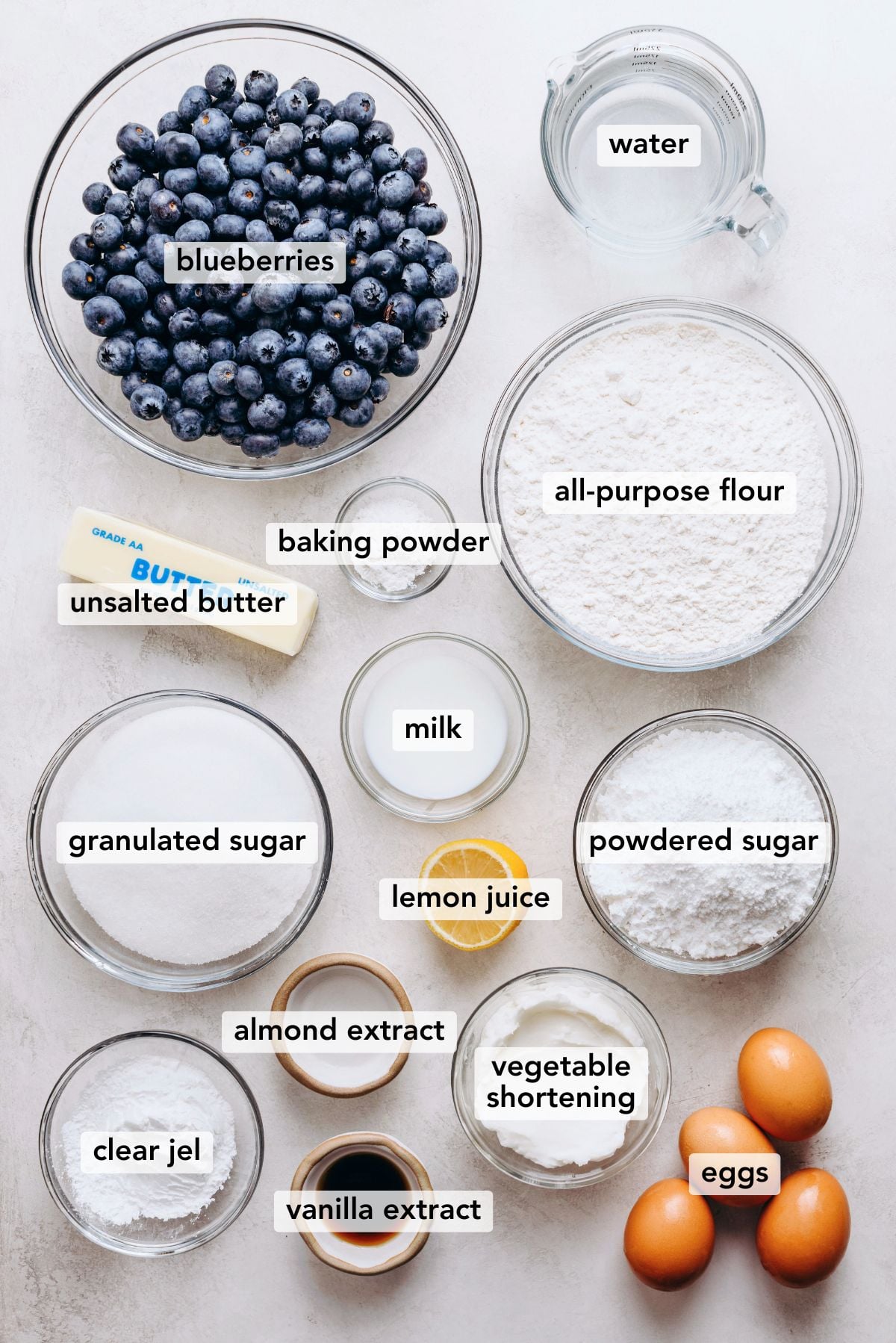 Ingredients in various size bowls and plates labeled as blueberries, water, unsalted butter, baking powder, granulated sugar, lemon juice, powdered sugar, clear jel, eggs, vanilla extract, and vegetable shortening, almond extract on a white marbled background.