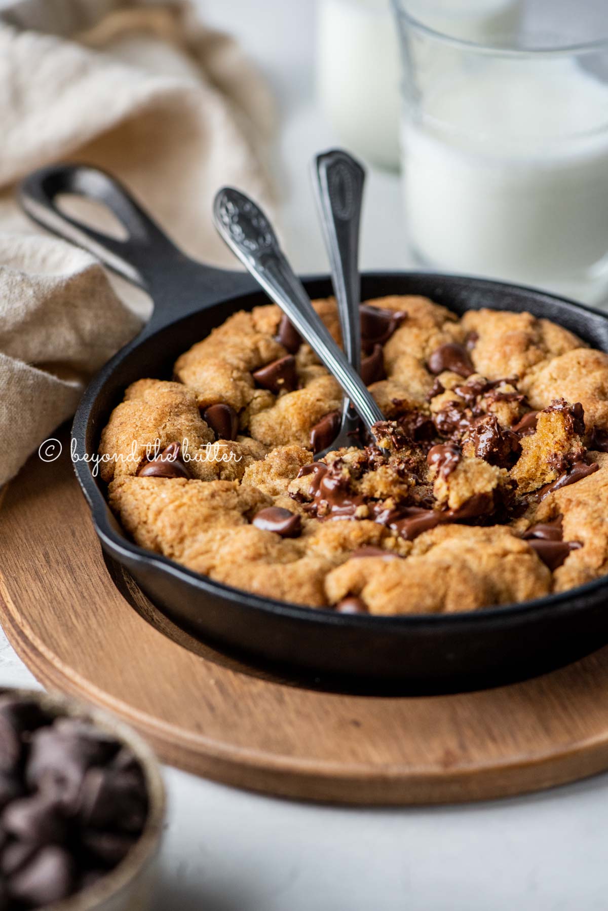 Warm brown butter chocolate chip skillet cookie with two spoons resting in skillet on wood trivet | All Images © Luyiz®