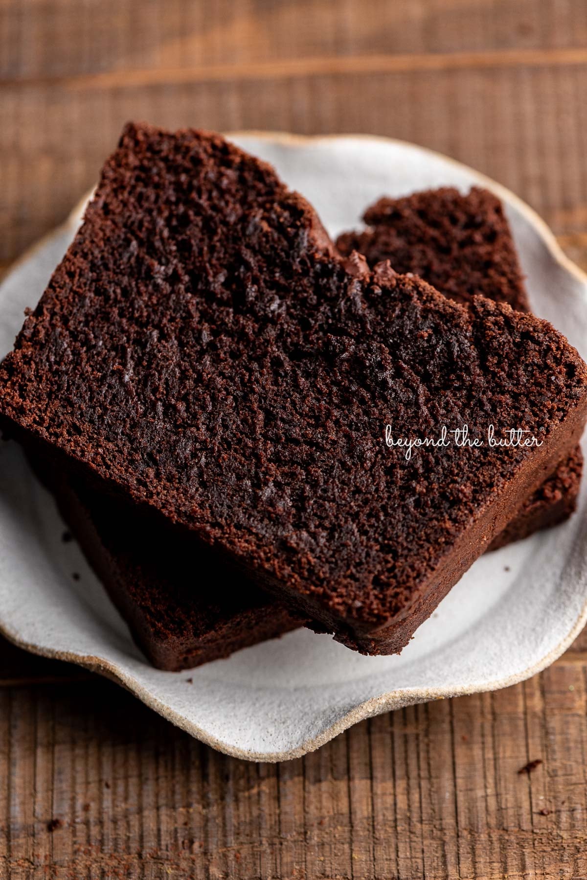Two thick slices of brownie bread on a cream colored, wavy edged ceramic dessert plate on a dark wood background.