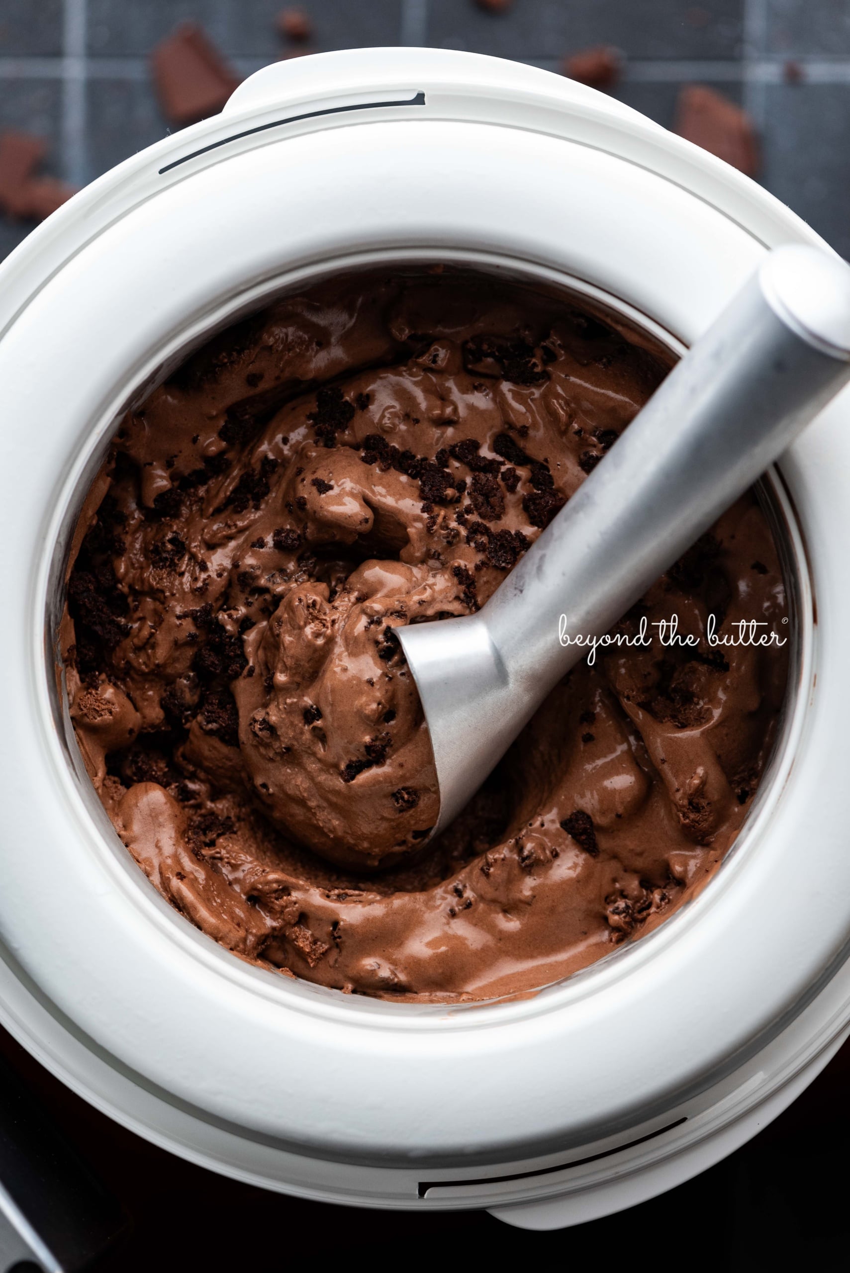 Scooping chocolate brownie ice cream out of the Ankarsrum ice cream maker attachment bowl with a chilled ice cream scoop on a dark background.