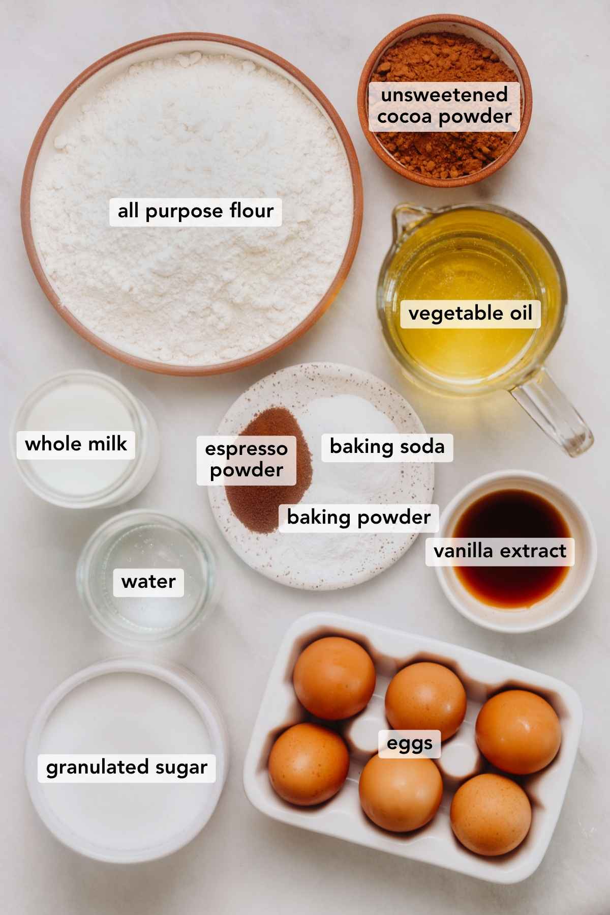 Ingredients in various size bowls labeled as all purpose flour, unsweetened cocoa powder, vegetable oil, whole milk, espresso powder, baking soda, baking powder, vanilla extract, water, eggs, and granulated sugar on a white marbled background.