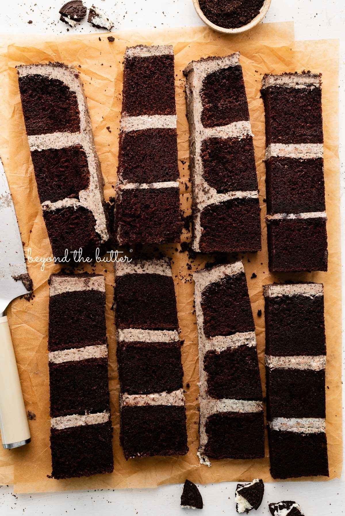 Slices of chocolate oreo cake on natural parchment paper with bits of oreo crumbs and a knife placed next to the cake.