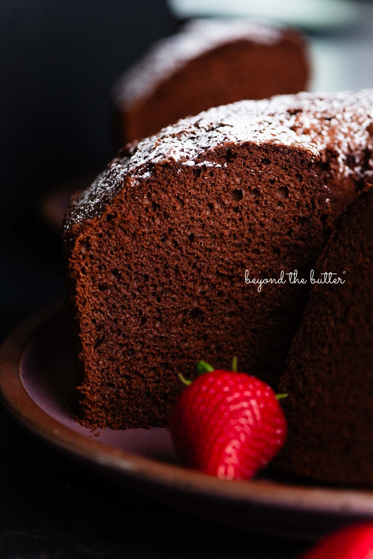 Chocolate chiffon cake sliced with whole strawberries placed on and in front of a dark purple ceramic plate on a black background.