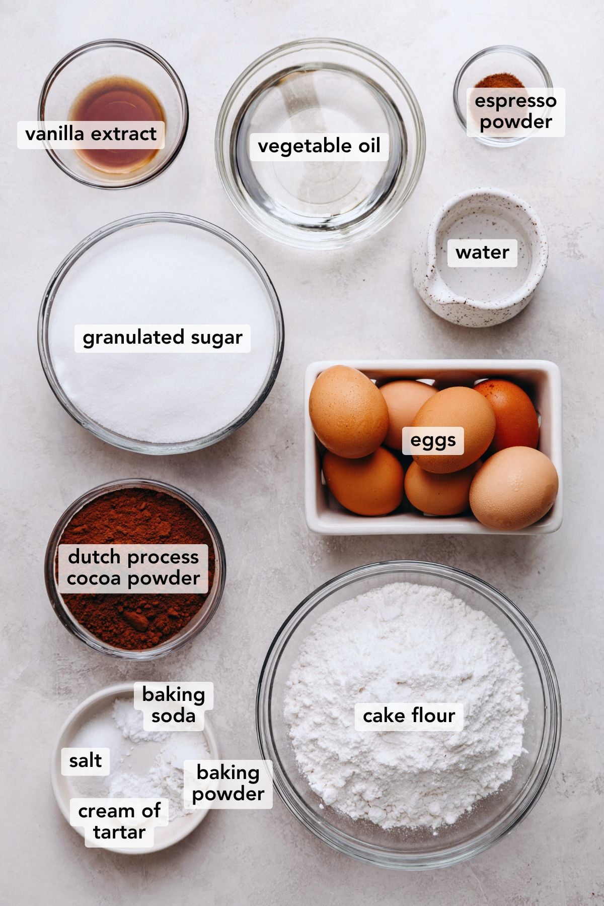 Ingredients in various size plates and bowls labeled as vanilla extract, vegetable oil, espresso powder, granulated sugar, water, eggs, dutch process cocoa powder, cake flour, baking soda, baking powder, cream of tartar, and salt on a white marbled background.