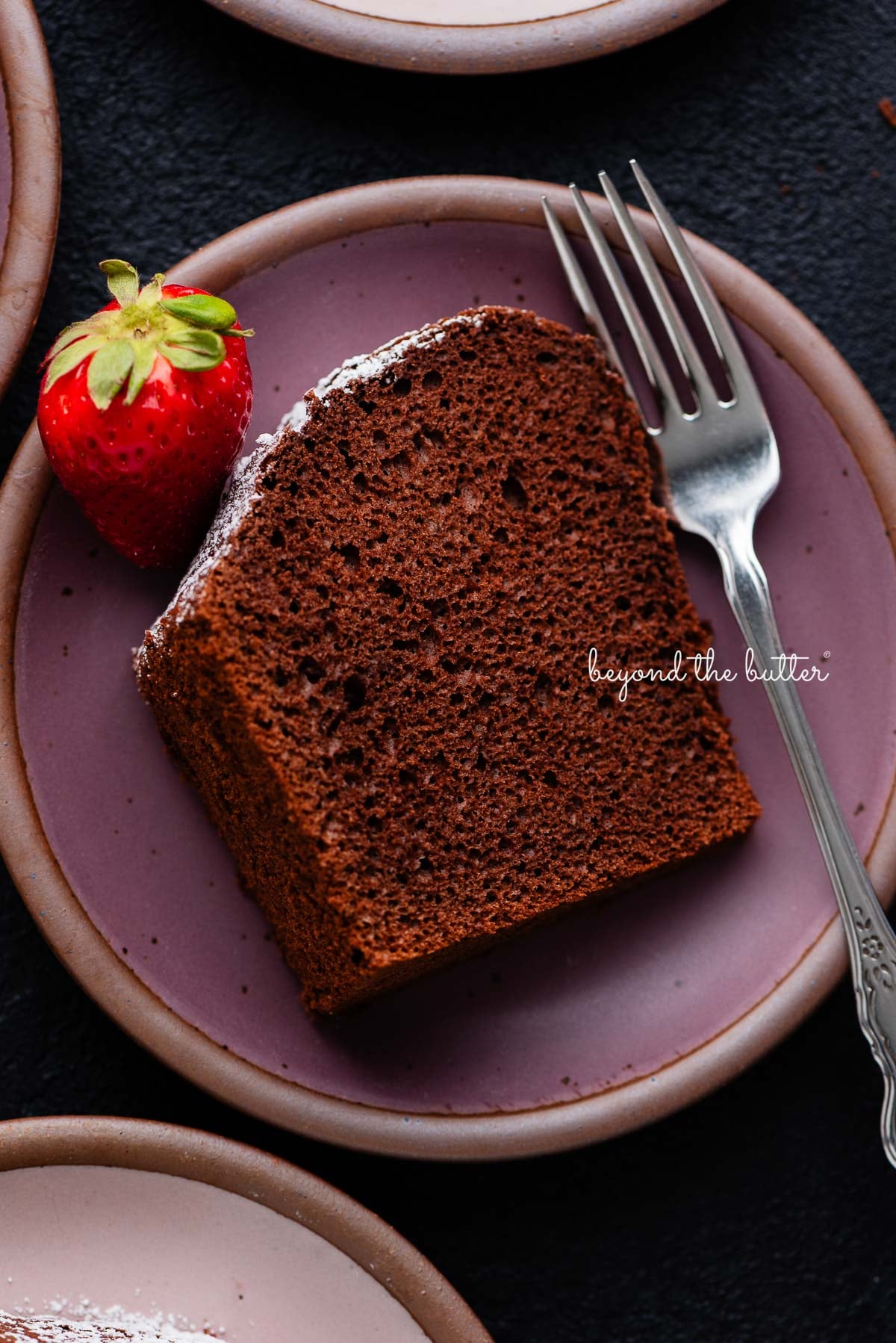 Slices of chocolate chiffon cake on small dark purple and light pink ceramic dessert plates with a fork and strawberry placed on each one on a black background.