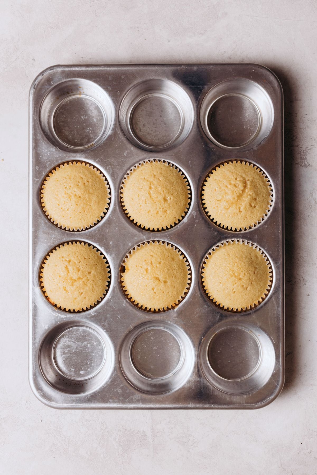 Six vanilla cupcakes baked in a metal cupcake tin on a white marbled background.