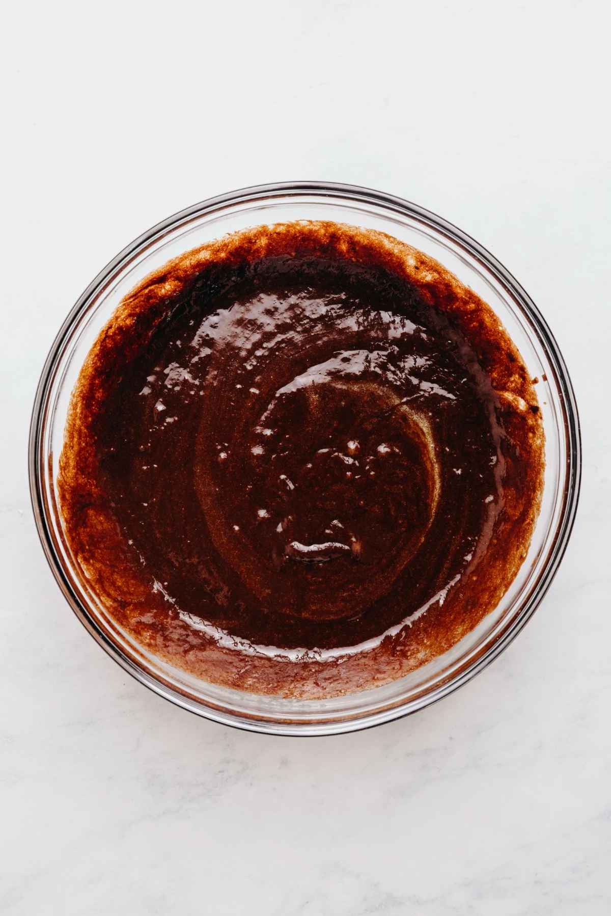 Whisked eggs, vanilla extract, granulated sugar, and melted chocolate mixture in a large glass mixing bowl on a white and grey marbled background.