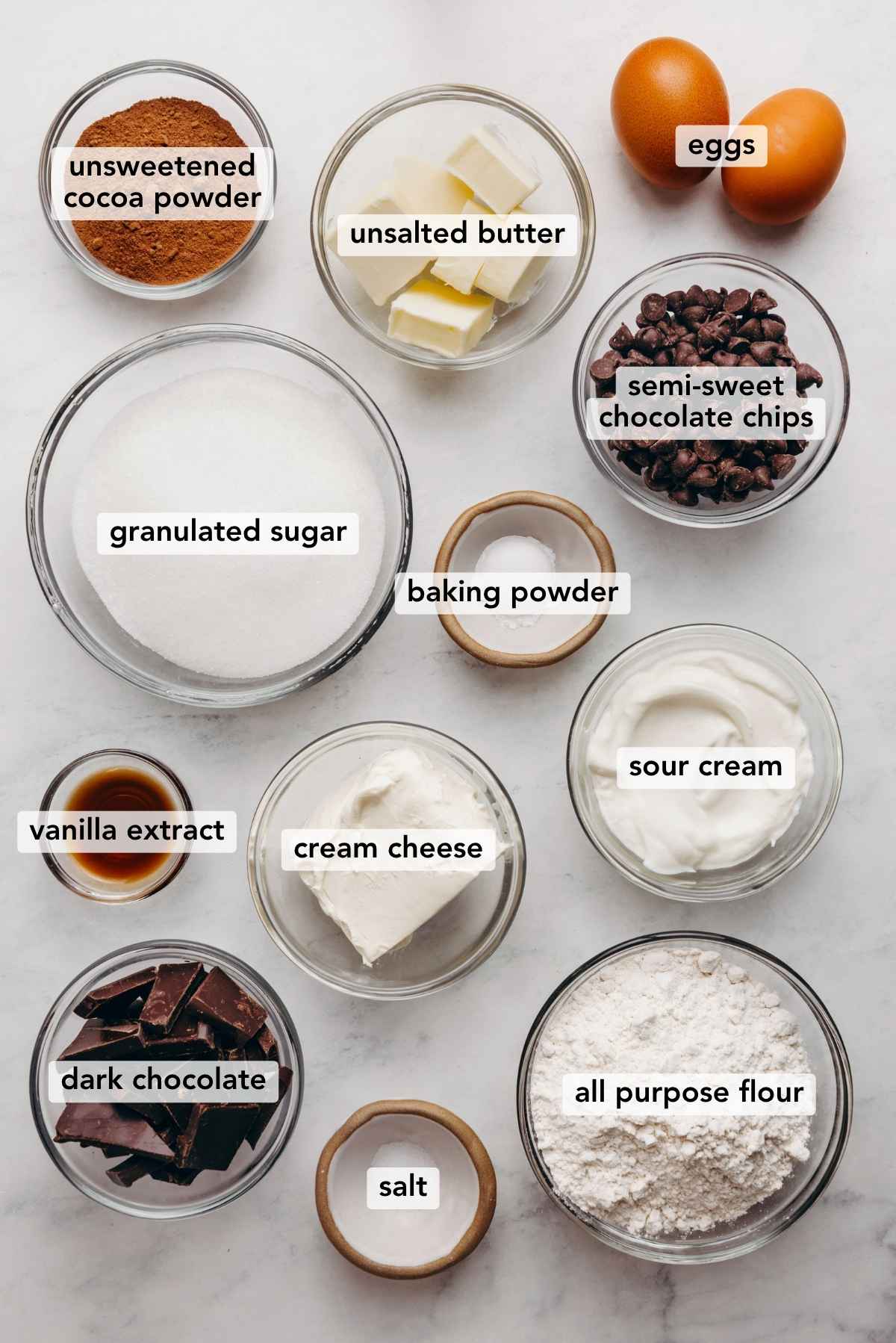 Ingredients in various size bowls labeled as unsweetened cocoa powder, unsalted butter eggs, granulated sugar, baking powder, semi sweet chocolate chips, vanilla extract, cream cheese, sour cream, dark chocolate, salt, and all purpose flour on a white marbled background.