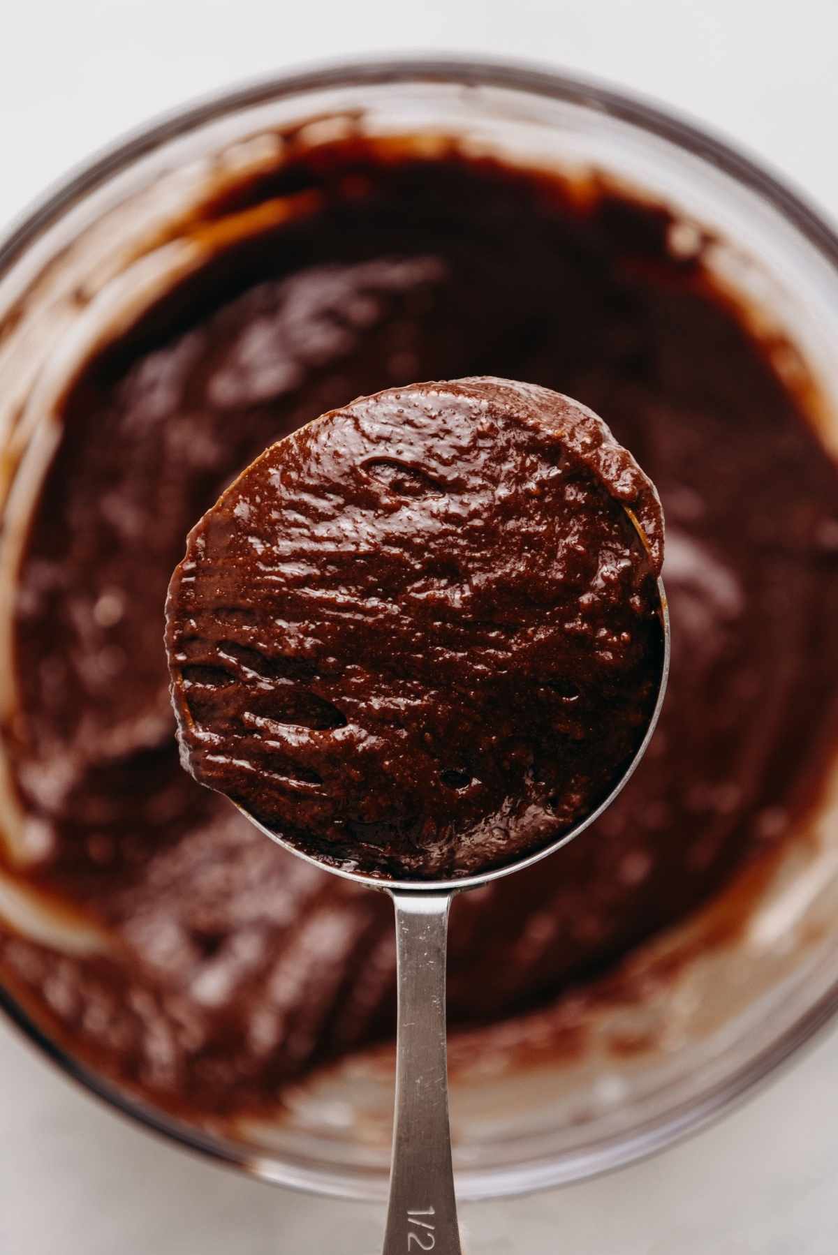 Brownie batter in a large glass mixing bowl with a one half cup measuring cup holding some reserved brownie batter.