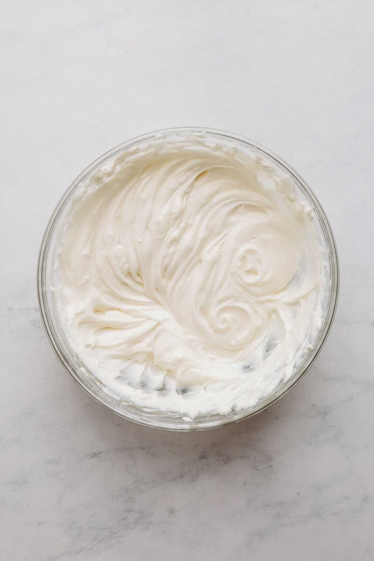Cream cheese, sour cream, and sugar mixed together in a large glass mixing bowl on a white and grey marbled background.
