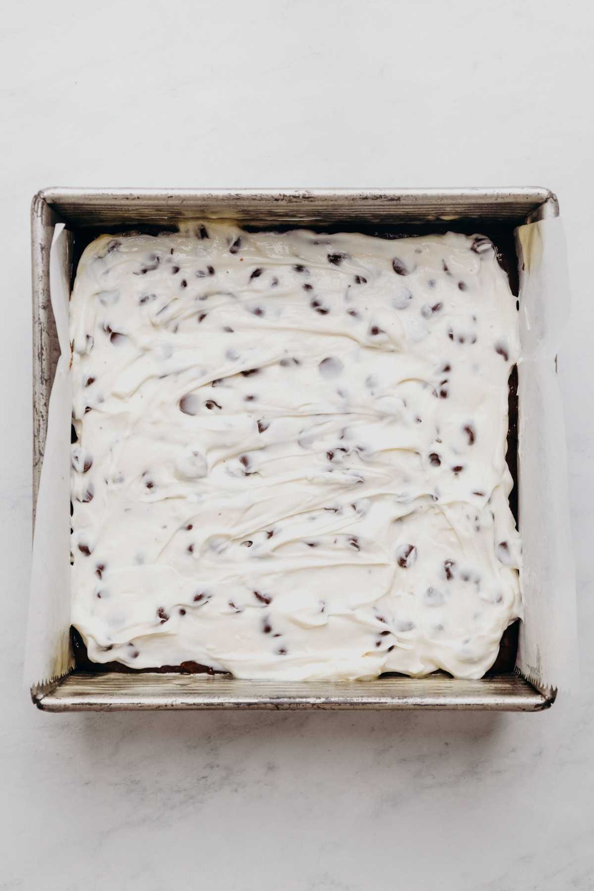 Brownie batter in a square metal baking tin lined with parchment paper with chocolate chip cream cheese filling on top.