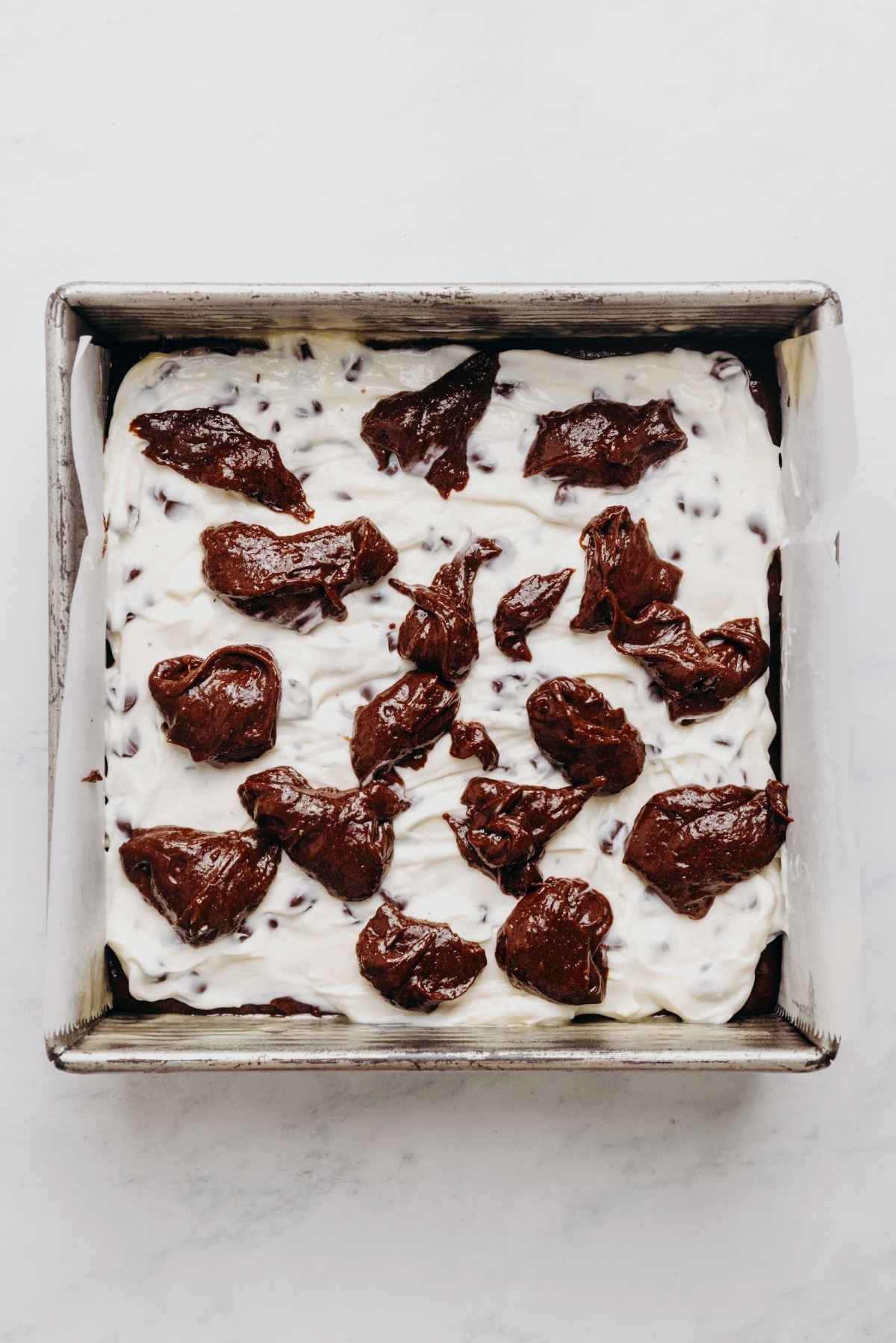 Dolloped brownie batter on top of chocolate chip cream cheese filling and brownie batter in a square parchment paper lined baking tin.