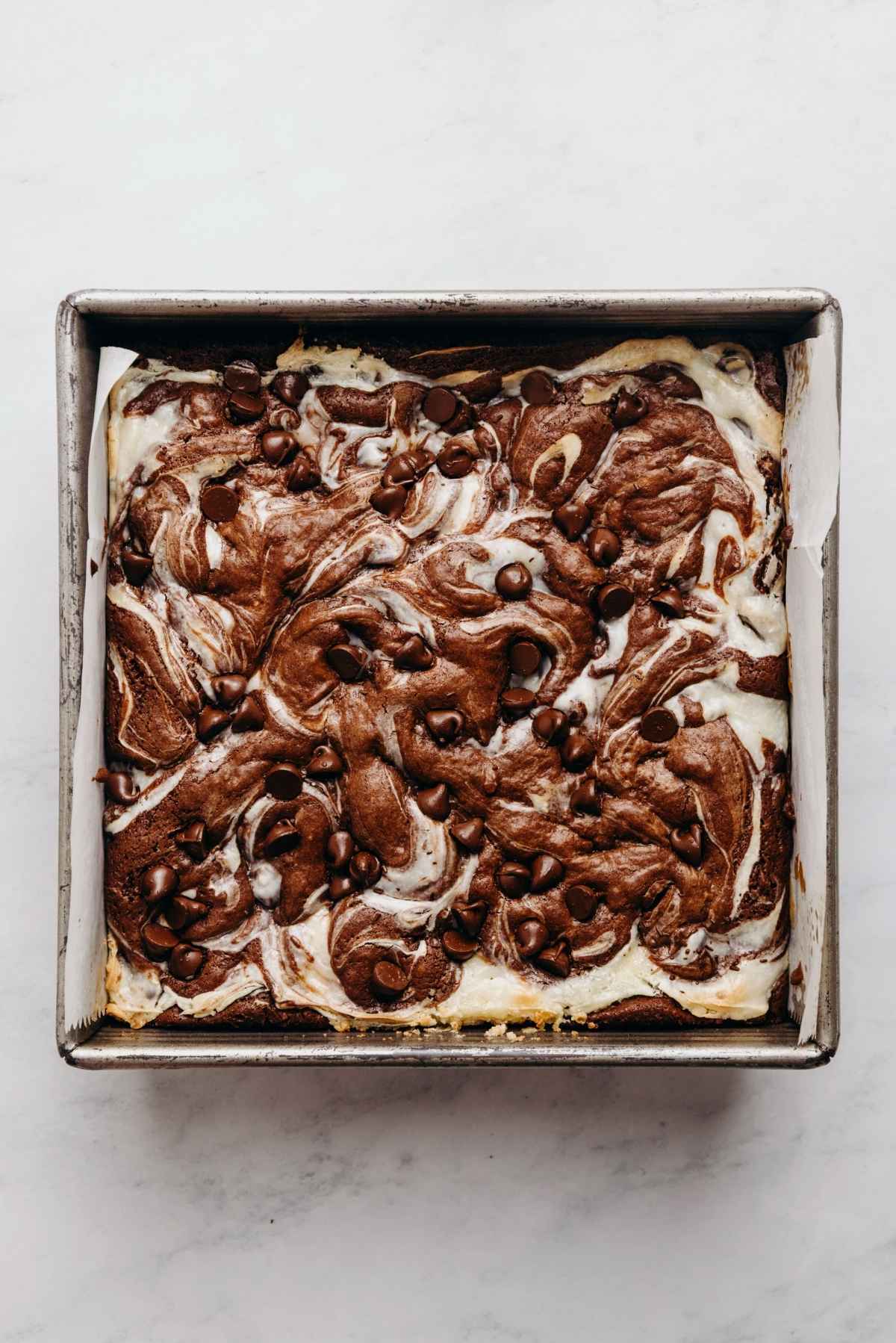 Baked chocolate chip cream cheese brownies in a parchment paper lined metal baking tin on a white and grey marbled background.