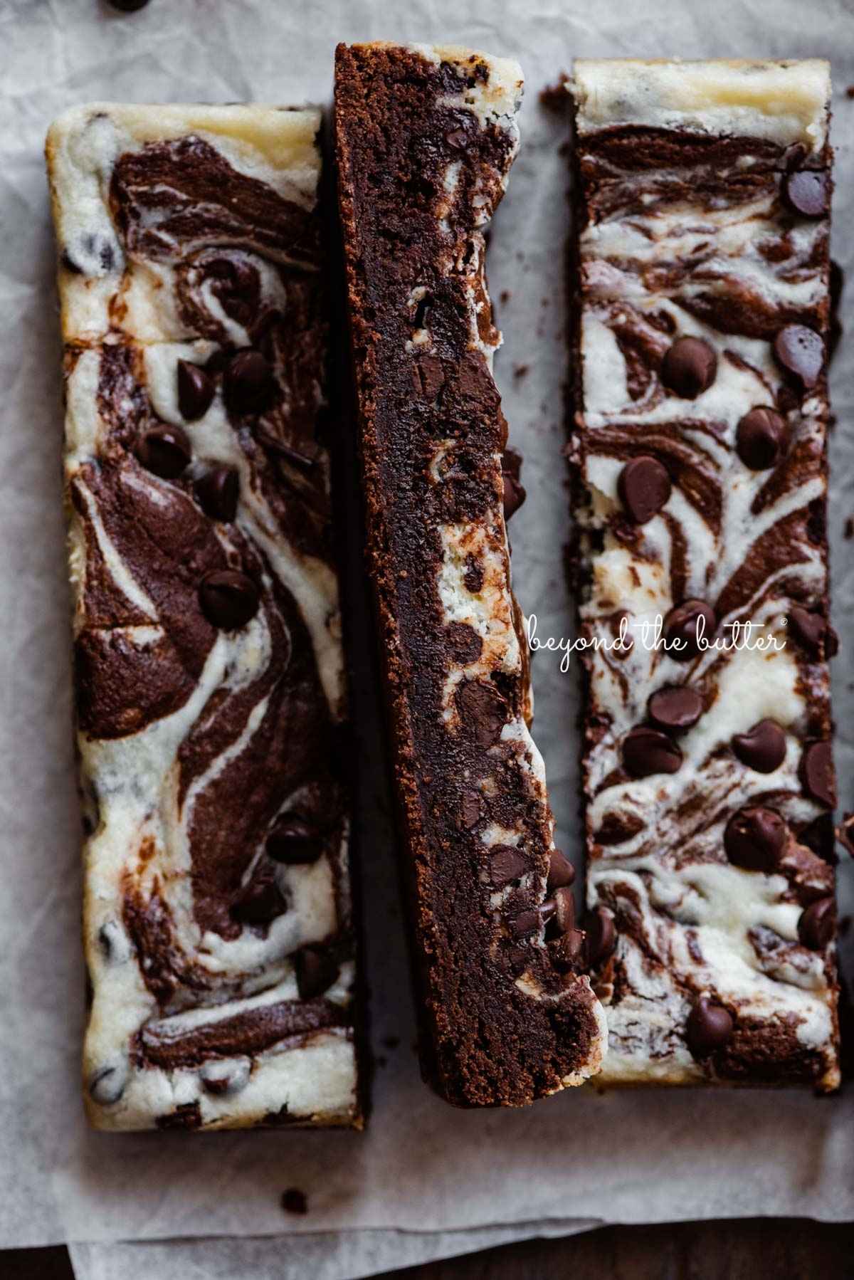 Chocolate chip cream cheese brownies cut into vertical bars on white parchment paper.