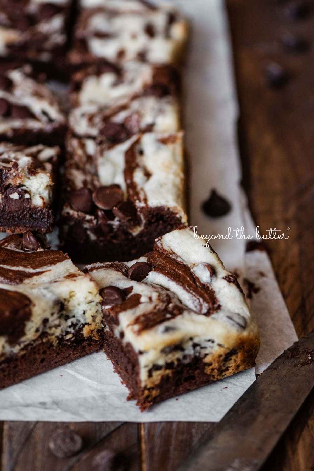 Chocolate chip cream cheese brownies on white parchment paper placed on a dark wood table.
