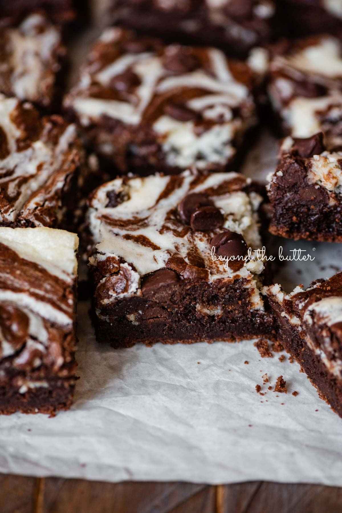 Sliced chocolate chip cream cheese brownies on white parchment paper lined placed on a dark wood background.