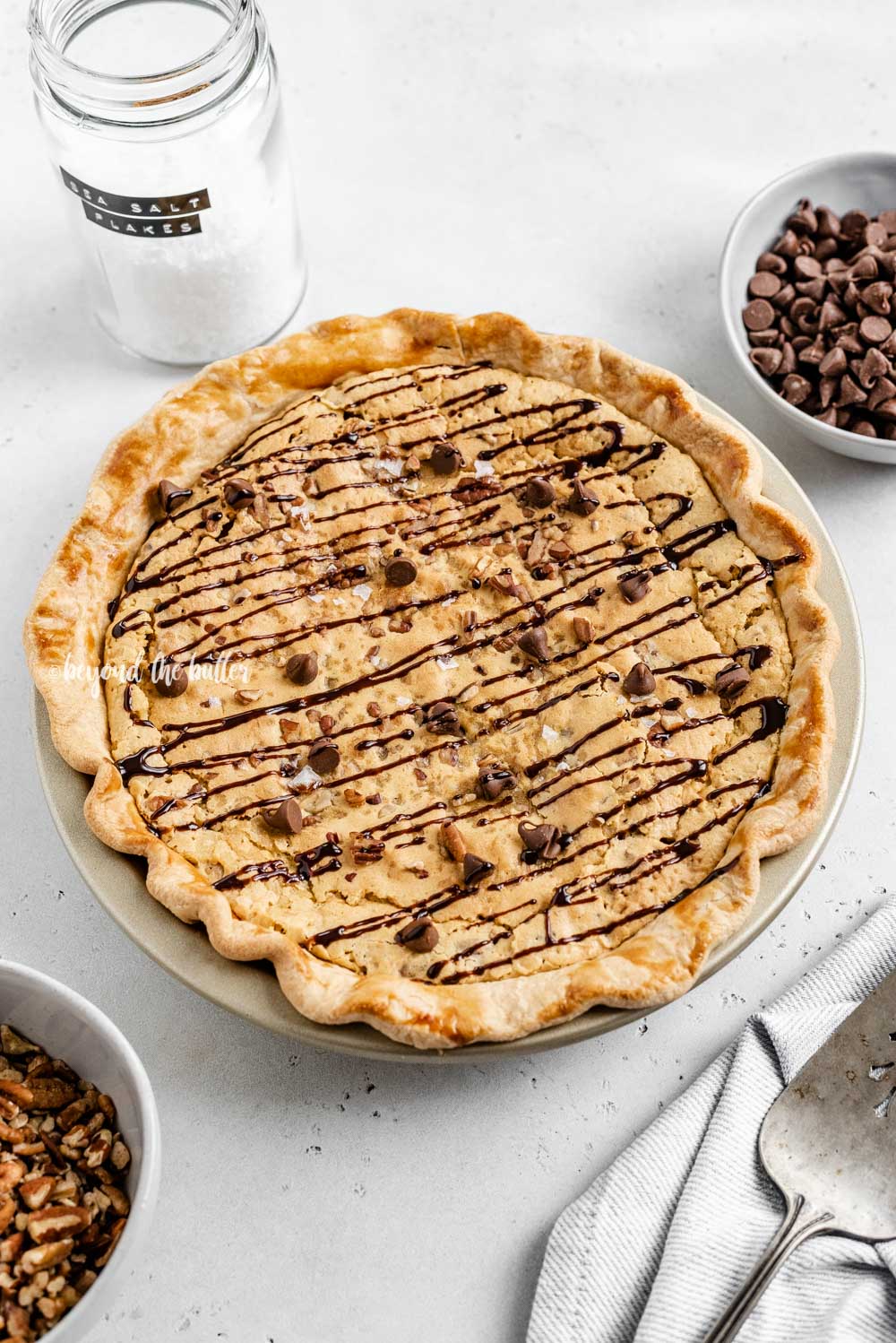 Overhead angled image of chocolate chip pie drizzled with chocolate syrup with bowls of chocolate chips, pecans, and a server | All Images © Luyiz™