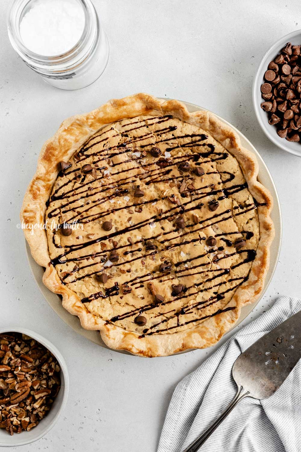 Overhead image of chocolate chip pie drizzled with chocolate syrup with bowls of chocolate chips, pecans, and a server | All Images © Luyiz™