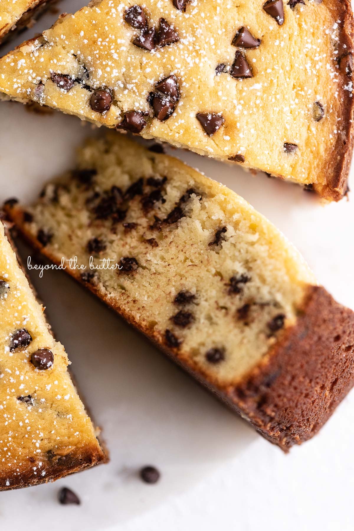 Slices of chocolate chip ricotta cake with one on its side on a marble cake round with small bowl of mini chocolate chips nearby | © Luyiz®