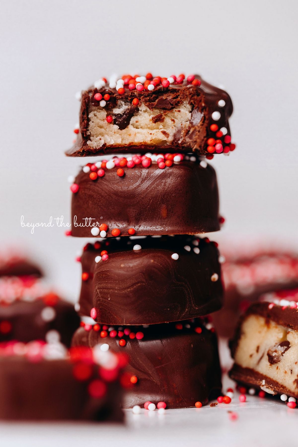 A stack of four chocolate covered cookie dough hearts topped with red, pink, and white sprinkles surrounded by other cookie dough hearts with a white background.