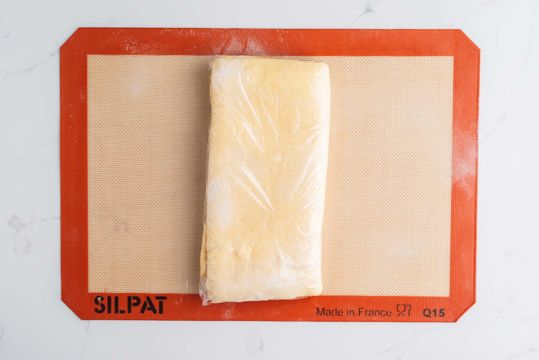 Laminated and folded croissant dough wrapped in plastic wrap on a silicone baking sheet.