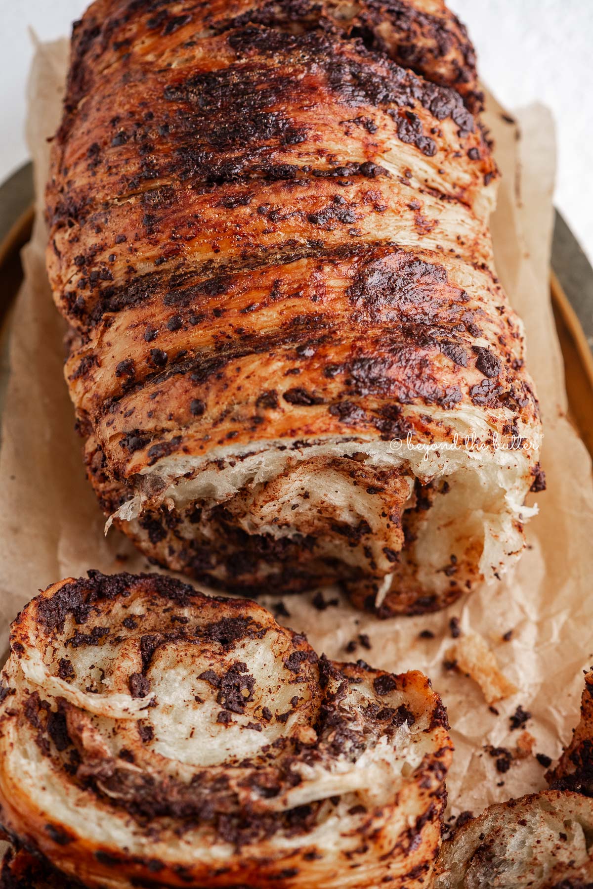 Sliced chocolate croissant bread loaf on brown parchment paper lined gold tray and white marbled background.