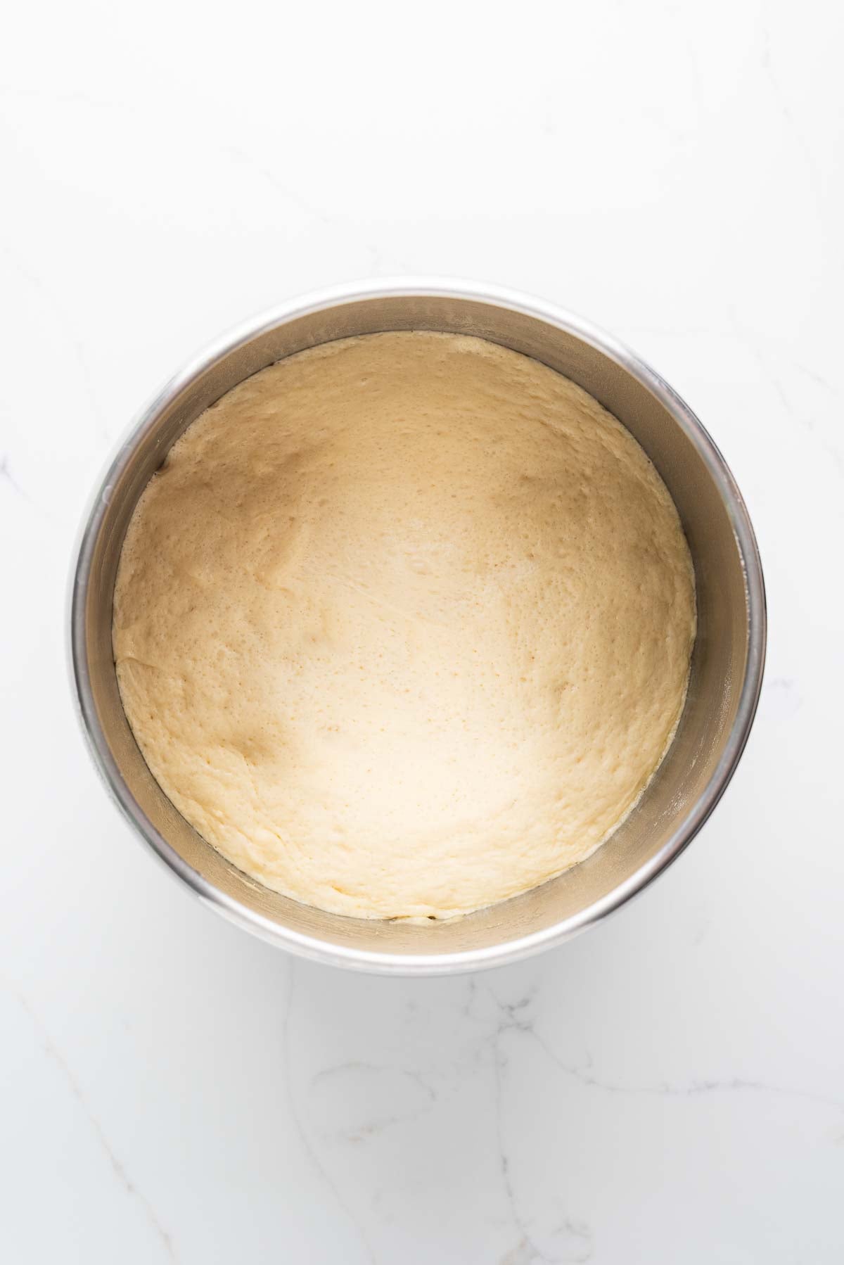 First rise of croissant dough in a stainless steel mixing bowl on a white marbled background.