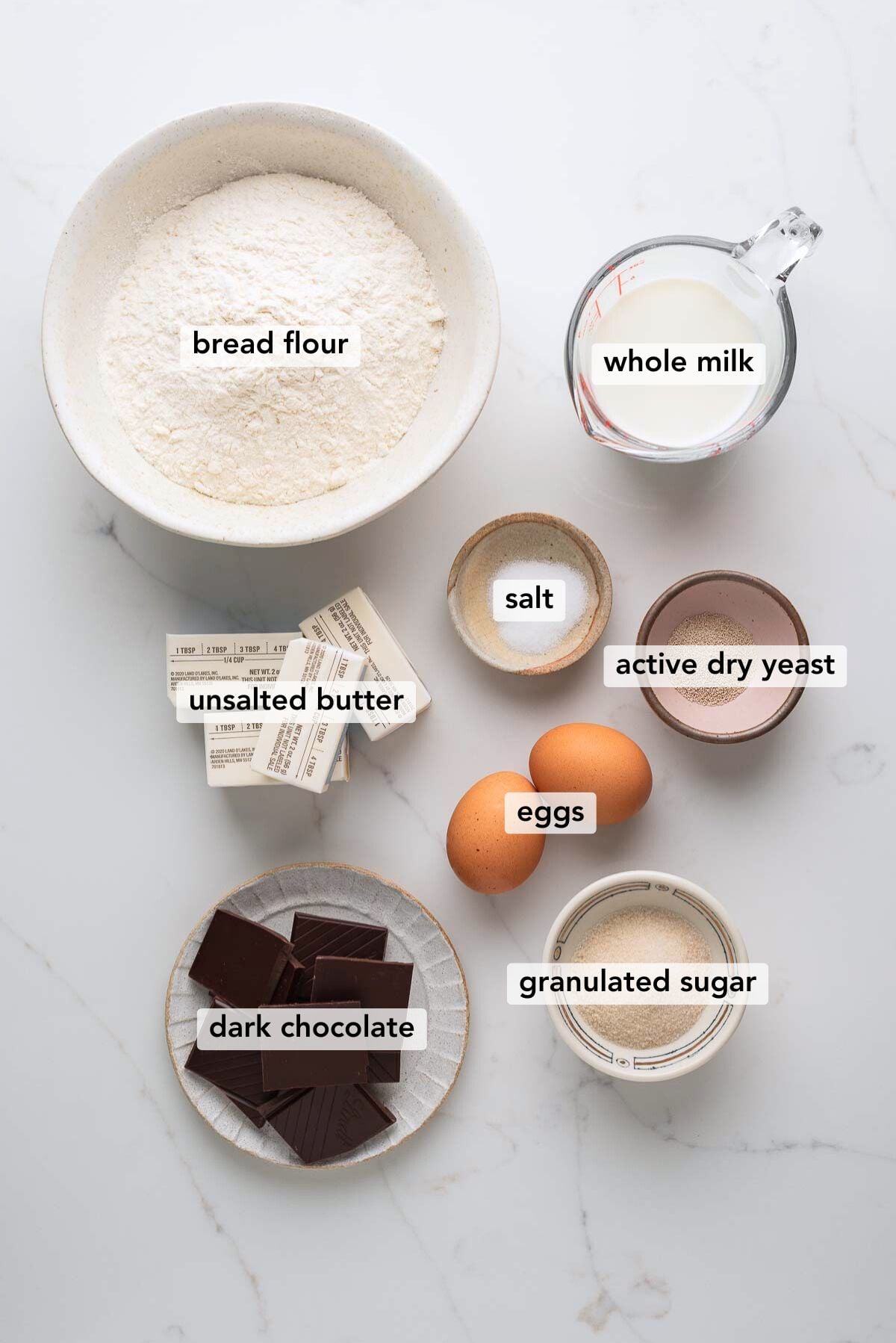Ingredients in various size bowls labeled as bread flour, whole milk, unsalted butter, salt, active dry yeast, eggs, dark chocolate, and granulated sugar on a white marbled background.
