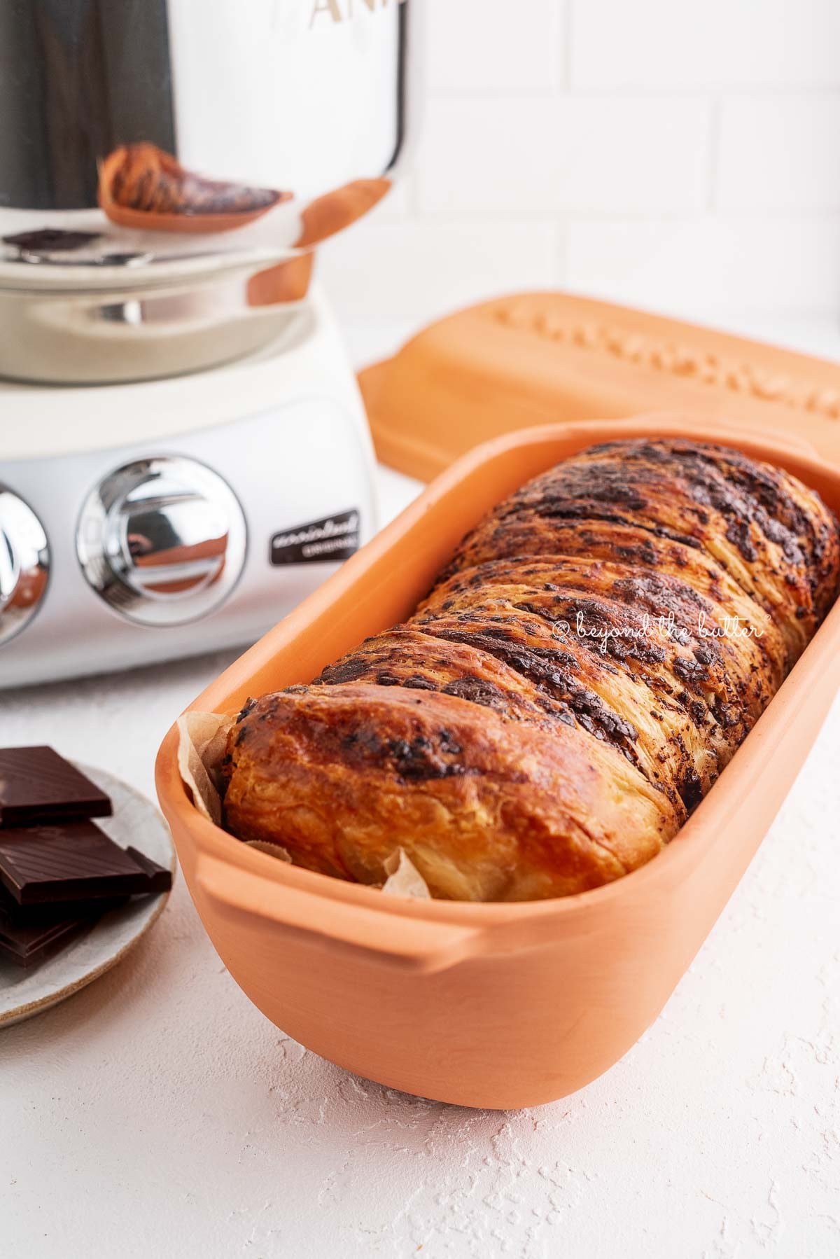 A chocolate croissant loaf placed in a parchment paper lined Ankasrum Clay Baker and next to a plate of dark chocolate and a cream colored Ankarsrum Assistent Original Mixer on a white background.