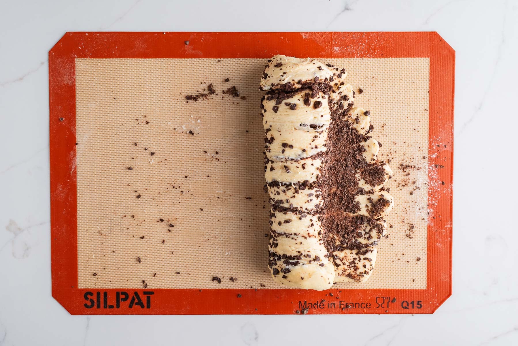 Rolling up the chocolate croissant dough loaf on a silicone baking sheet on a white marbled background.