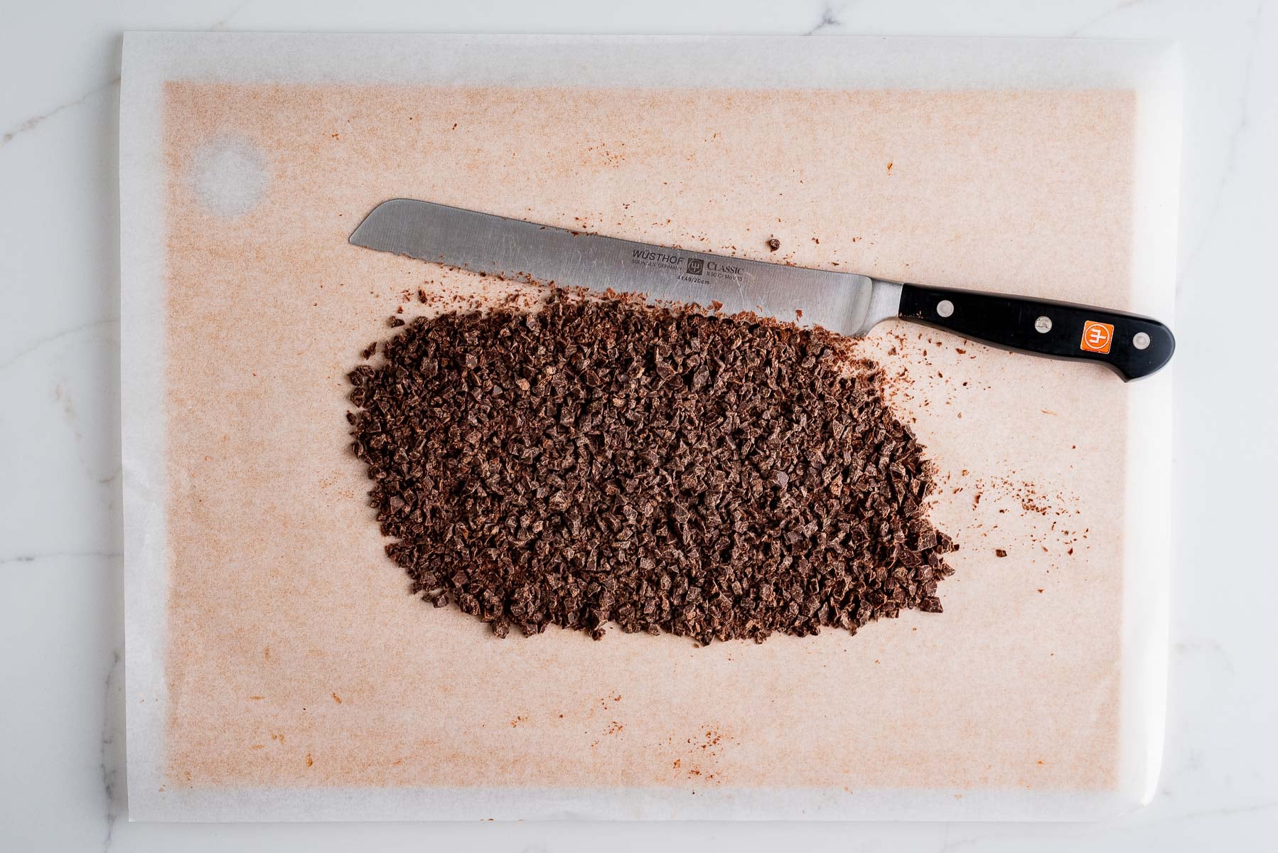 Finely chopped dark chocolate on a parchment paper lined cutting board with a serrated knife on a white marbled background.