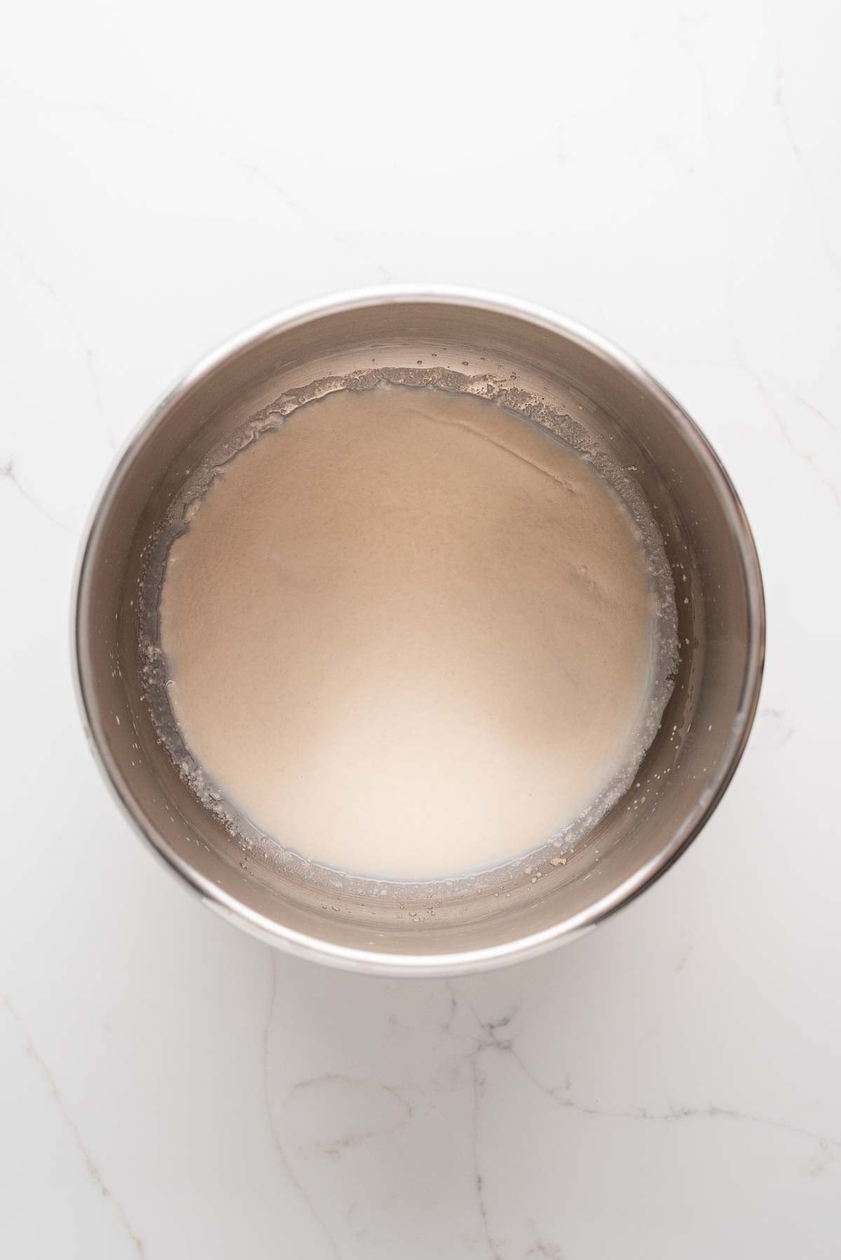Proofing yeast in warm milk and sugar in a large stainless steel Ankarsrum mixing bowl on a white marbled background.