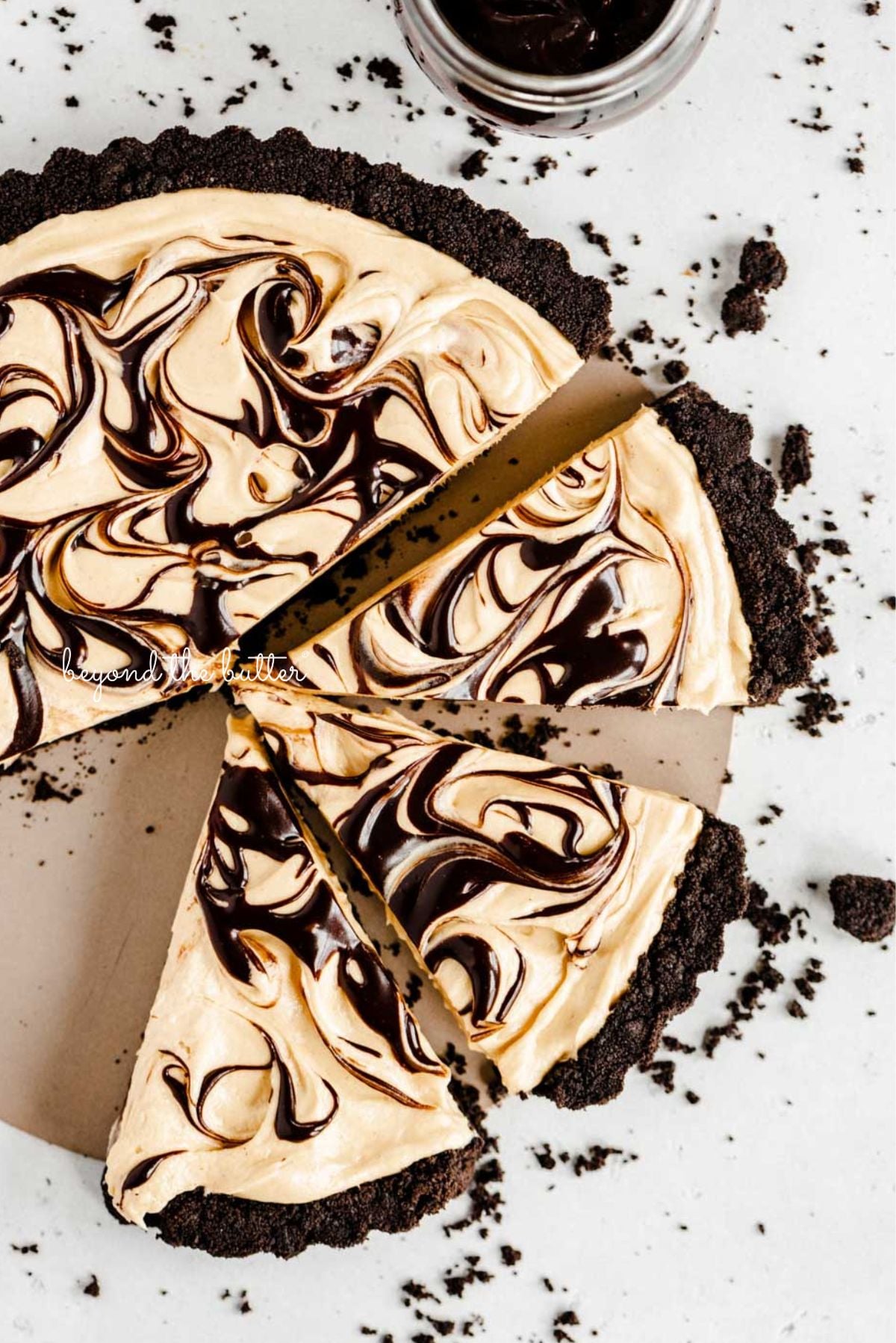 Round chocolate peanut butter tart swirled with chocolate syrup and cut into slices with a bowl of chocolate sandwich crumbs next to it on a white speckled background.