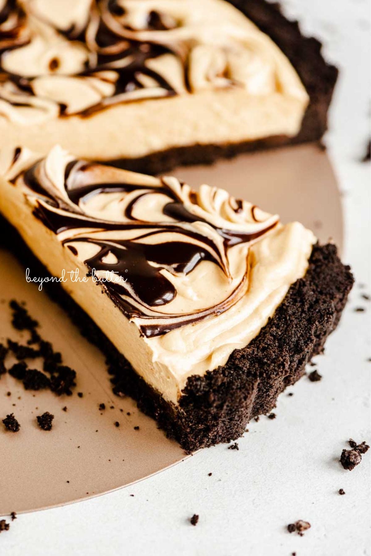 Sliced chocolate peanut butter tart swirled with chocolate syrup surrounded by a few chocolate sandwich cookie crumbs on a white speckled background.